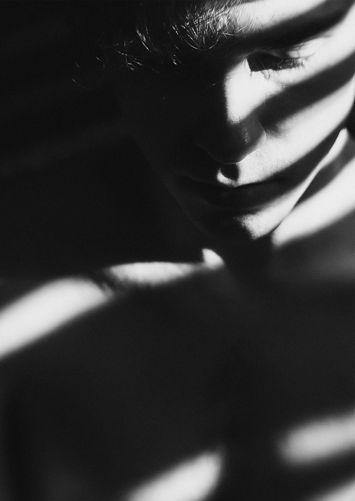 A young, shirtless boy in a darkened room with light from slightly opened blinds looks down. His face and upper torso are awash in shadow and light.