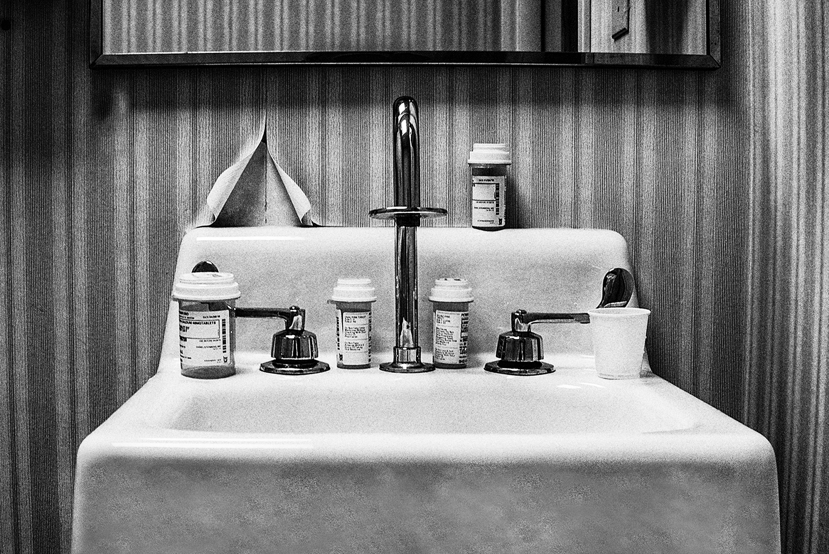 Close-up of the top of a small, white, pedestal bathroom sink with silver fixtures and the bottom part of a rectangular mirror against peeling, striped wallpaper. On the sink are pill bottles and a small plastic cup.