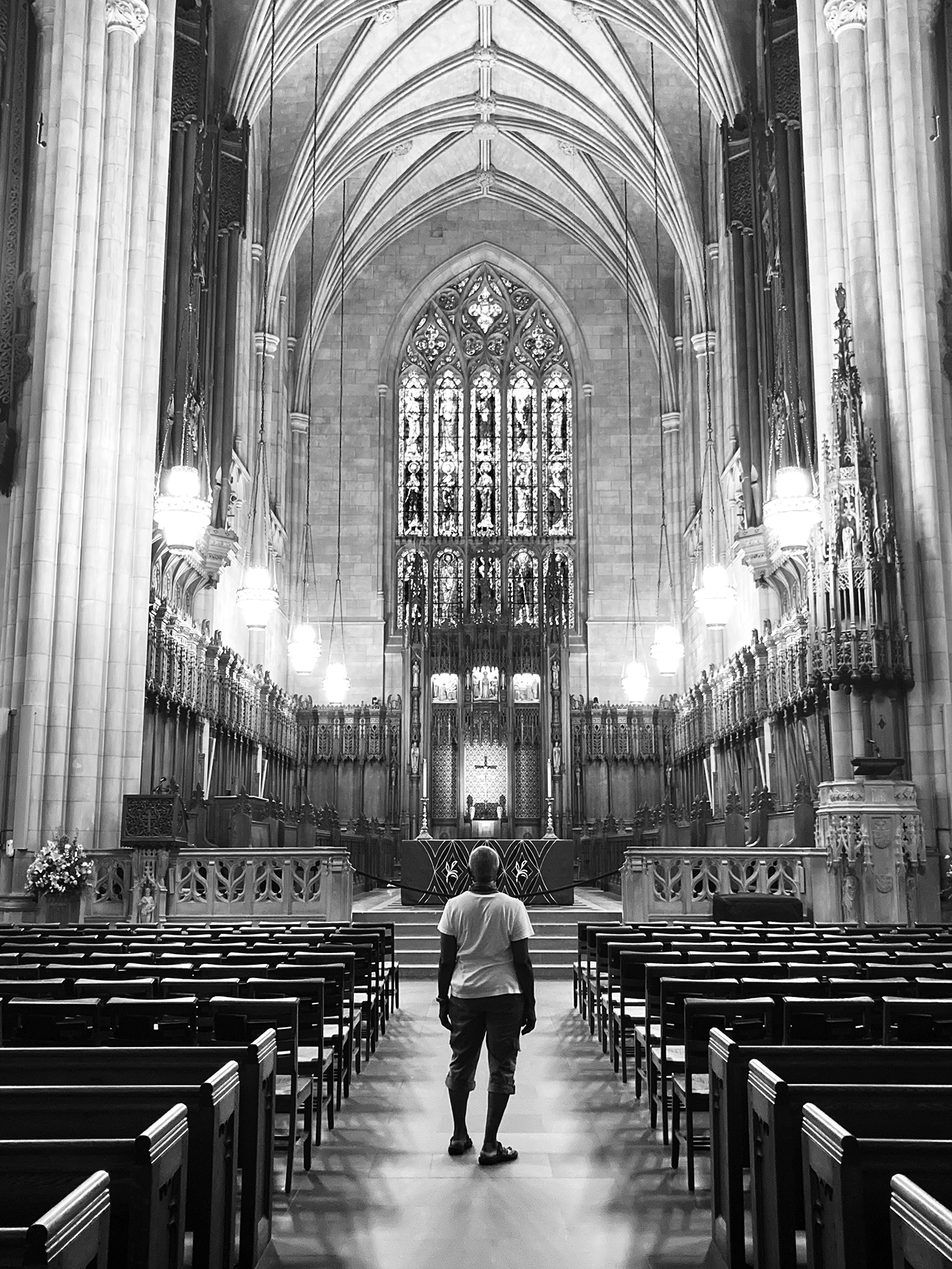 Interior of a large, impressive looking church with a high, rib-vaulted ceiling, tall columns, and a large stained-glass feature on the back wall. A casually dressed person stands in the center aisle and looks toward the back wall.