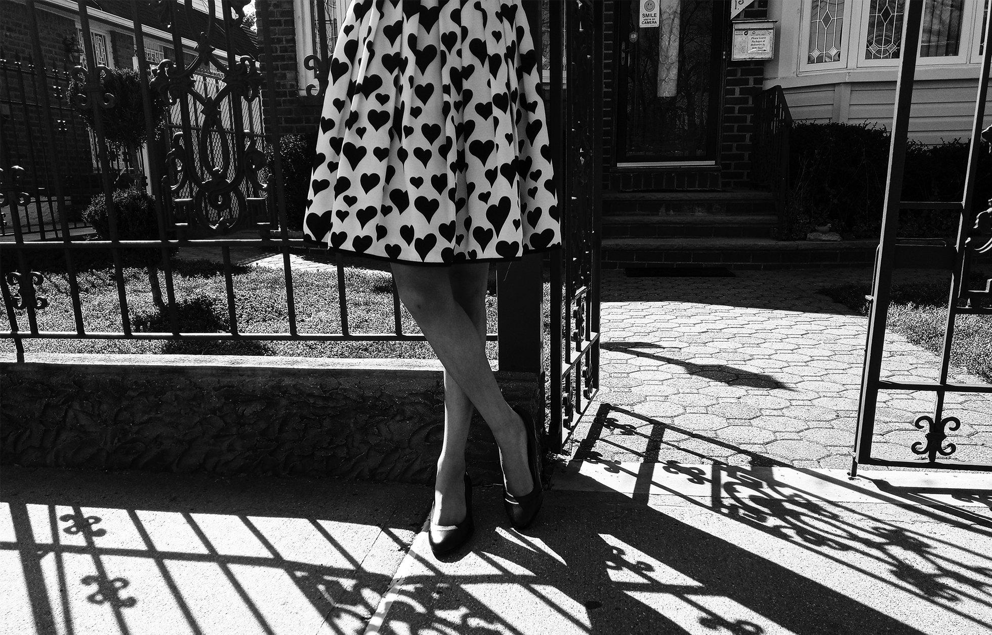 A woman stands with her legs crossed near a wrought-iron gate in front of a house. She is shown from the waist down. She is wearing a white dress with hearts all over it and a pair of pumps.