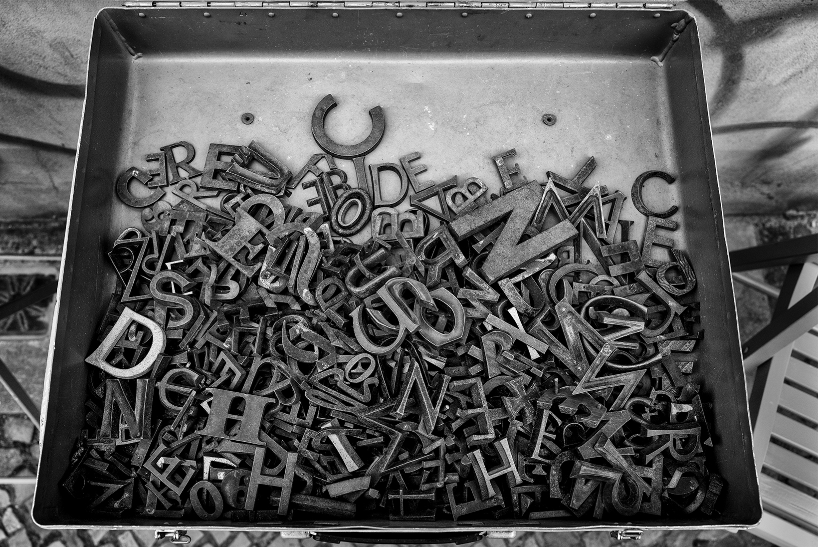 The bottom part of a hinged metal box half full of metal letters of various sizes.
