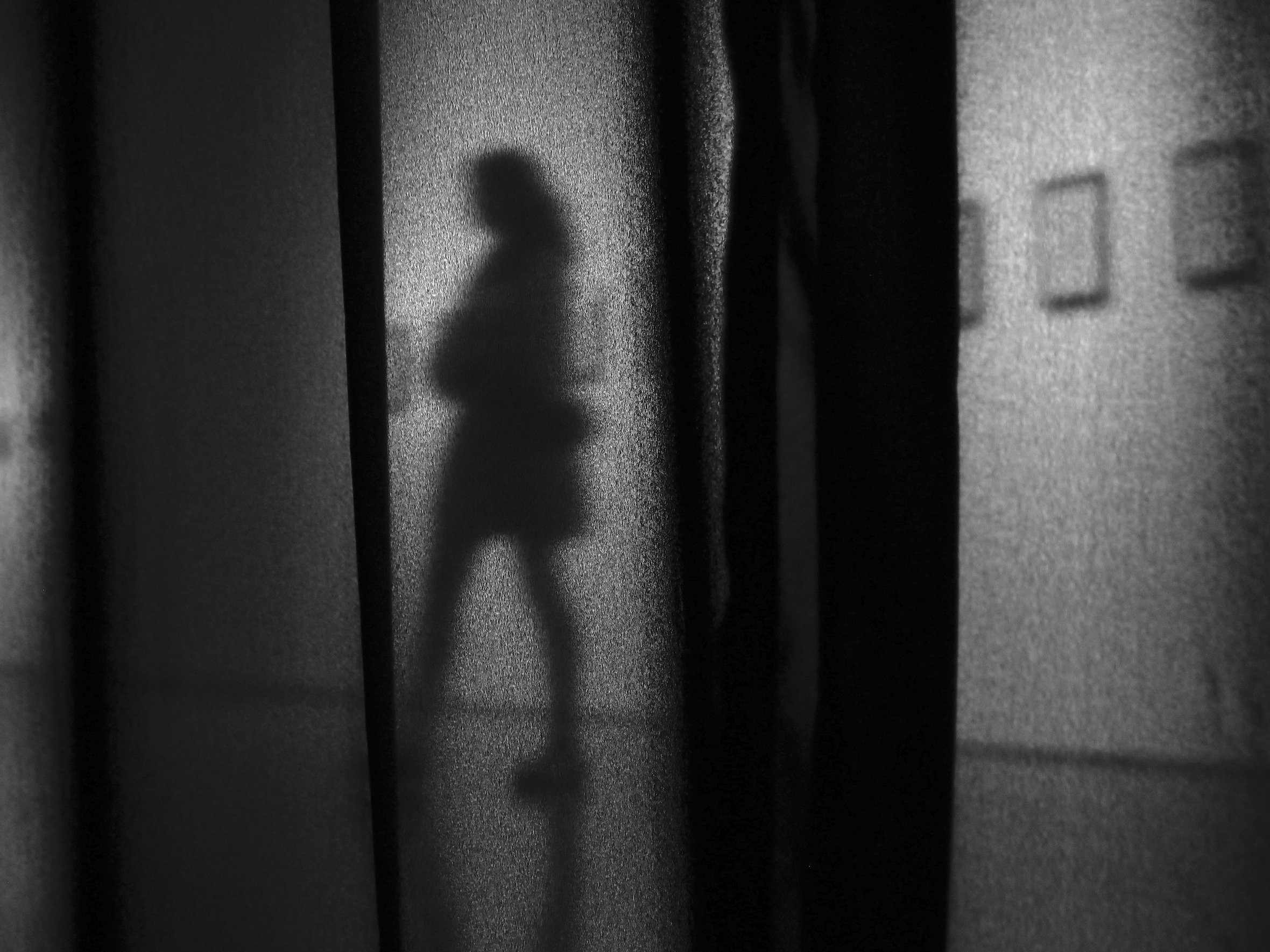 The silhouette of a girl in a skirt as she stands in a room with many same-size, evenly-spaced, square objects on the wall behind her seen through a gauzy curtain.