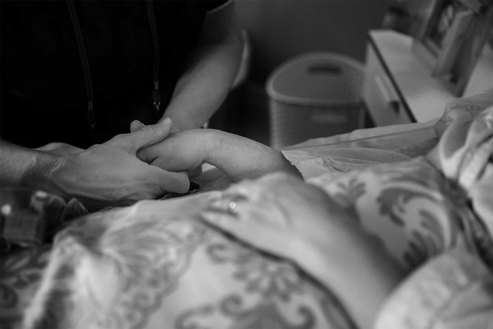 A person stands at a bedside and holds the right hand of the woman who is in bed. The focus of the image is on the hand-holding: the faces of the people are not visible.