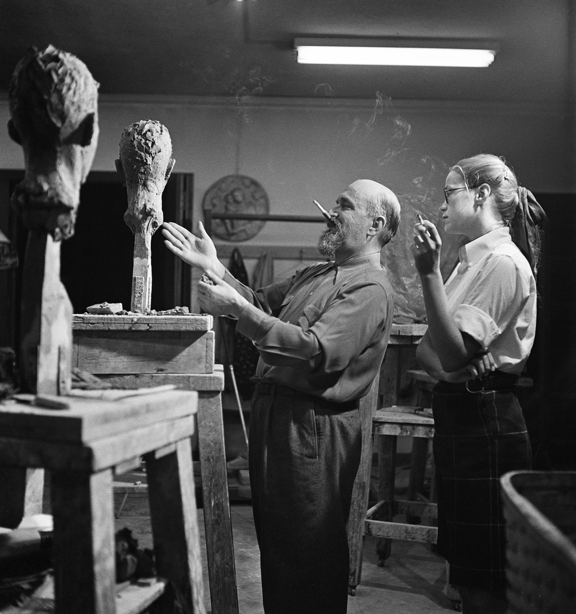 An older man and younger woman stand in an art studio or classroom discussing one of the two clay busts on pedestals. Both people are smoking. The man has a cigarette in his mouth as he gestures to the bust with both hands.