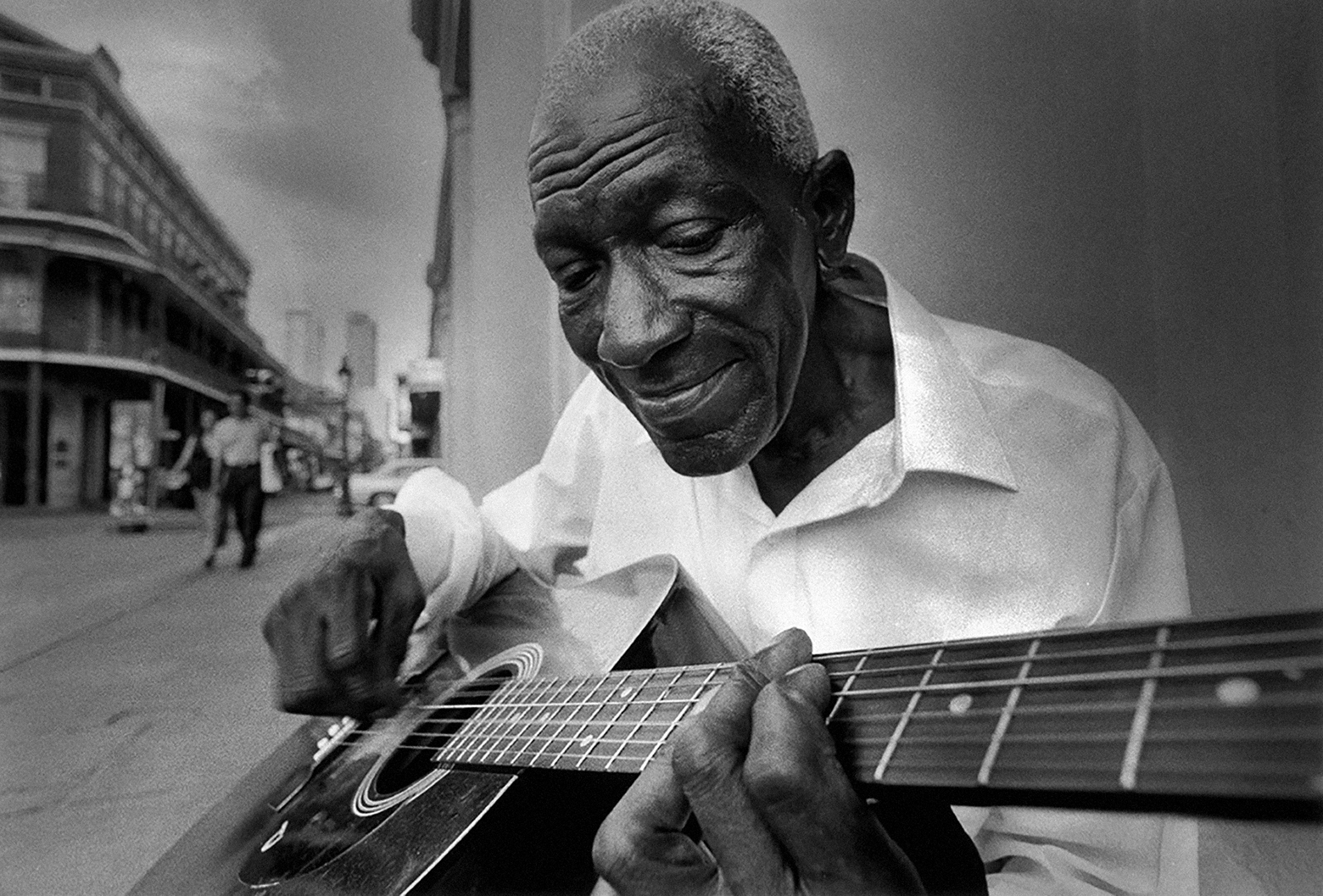 Close-up of a man who sits on a chair on the sidewalk and smiles as he looks down at the guitar he plays.