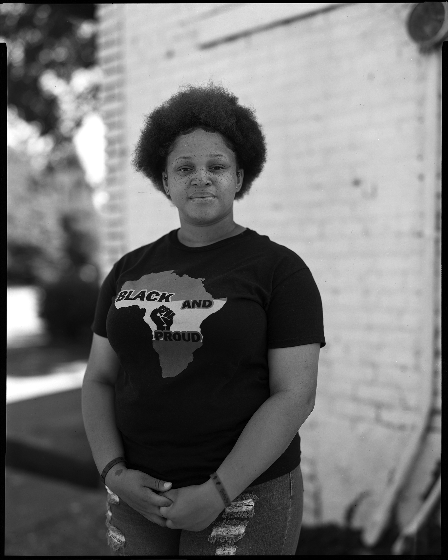 Aariona stands in front of a brick wall in Cleveland, Ohio, with her hands clasped in front of her. She looks into the camera with a pleasant, low-key, closed-mouth smile. Aariona has an Afro and wears a T-shirt that reads “Black and Proud.”