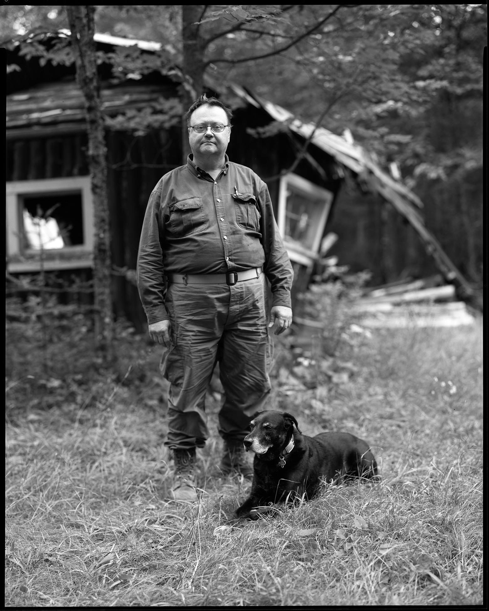 John looks into the camera as he stands outside of the cabin his father and grandfather took him to as a child in Two Harbors, Minnesota. A medium-size black dog is in the grass at his feet.
