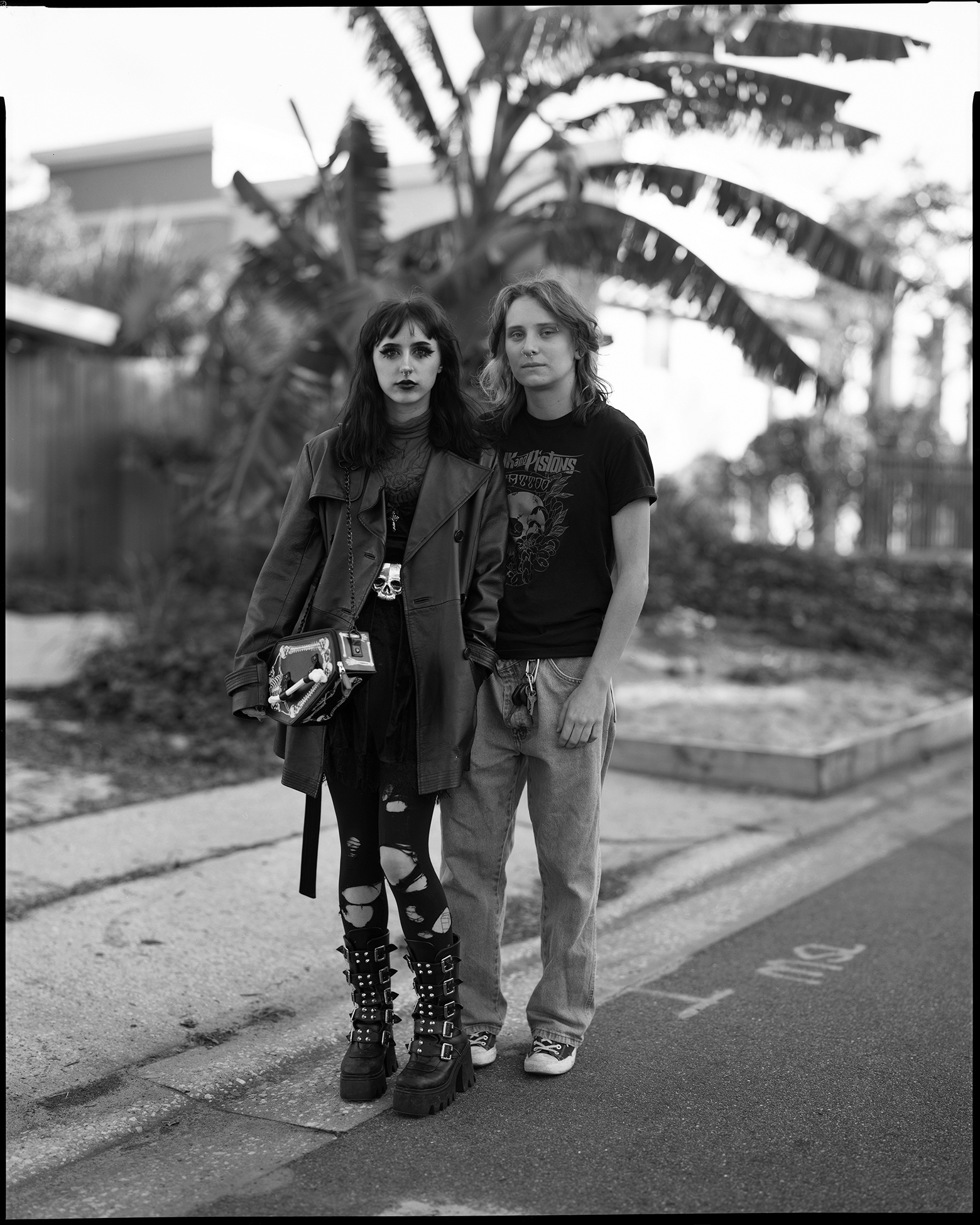 Juno and Peyton stand on a street next to the sidewalk in Maitland, Florida, and look into the camera. Juno is in front and slightly to the side of Peyton. Juno is wearing black leggings, with assorted holes, and thick, chunky, midcalf black boots.
