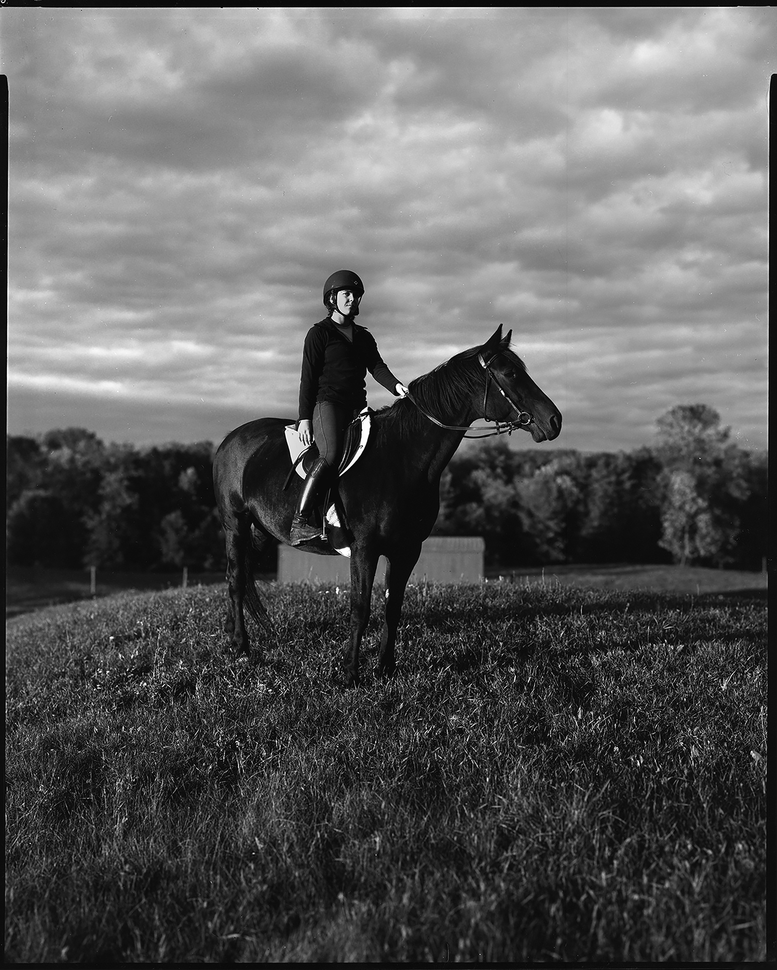 Saxon sits atop Peazie, a horse, in profile in a meadow in North Hero, Vermont, with many trees in the background on a cloudy day. Saxon wears riding boots and a helmet while looking toward the camera.