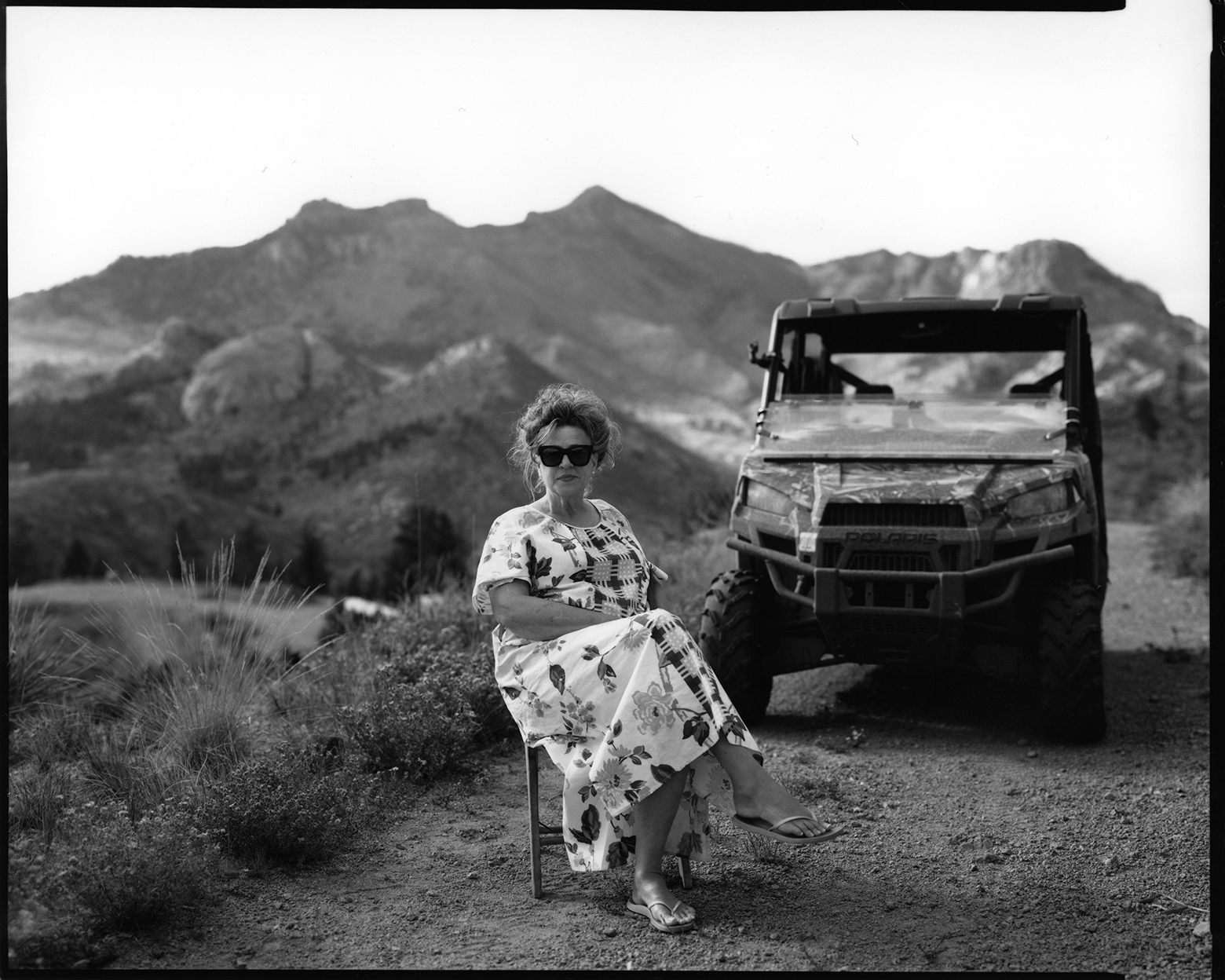 Tammy sits on a chair with her legs crossed on a dirt road in front of a jeep with hills in the background in Westcreek, Colorado. Tammy has on dark glasses as she looks toward the camera wearing a long, white, flower-print dress and sandals.