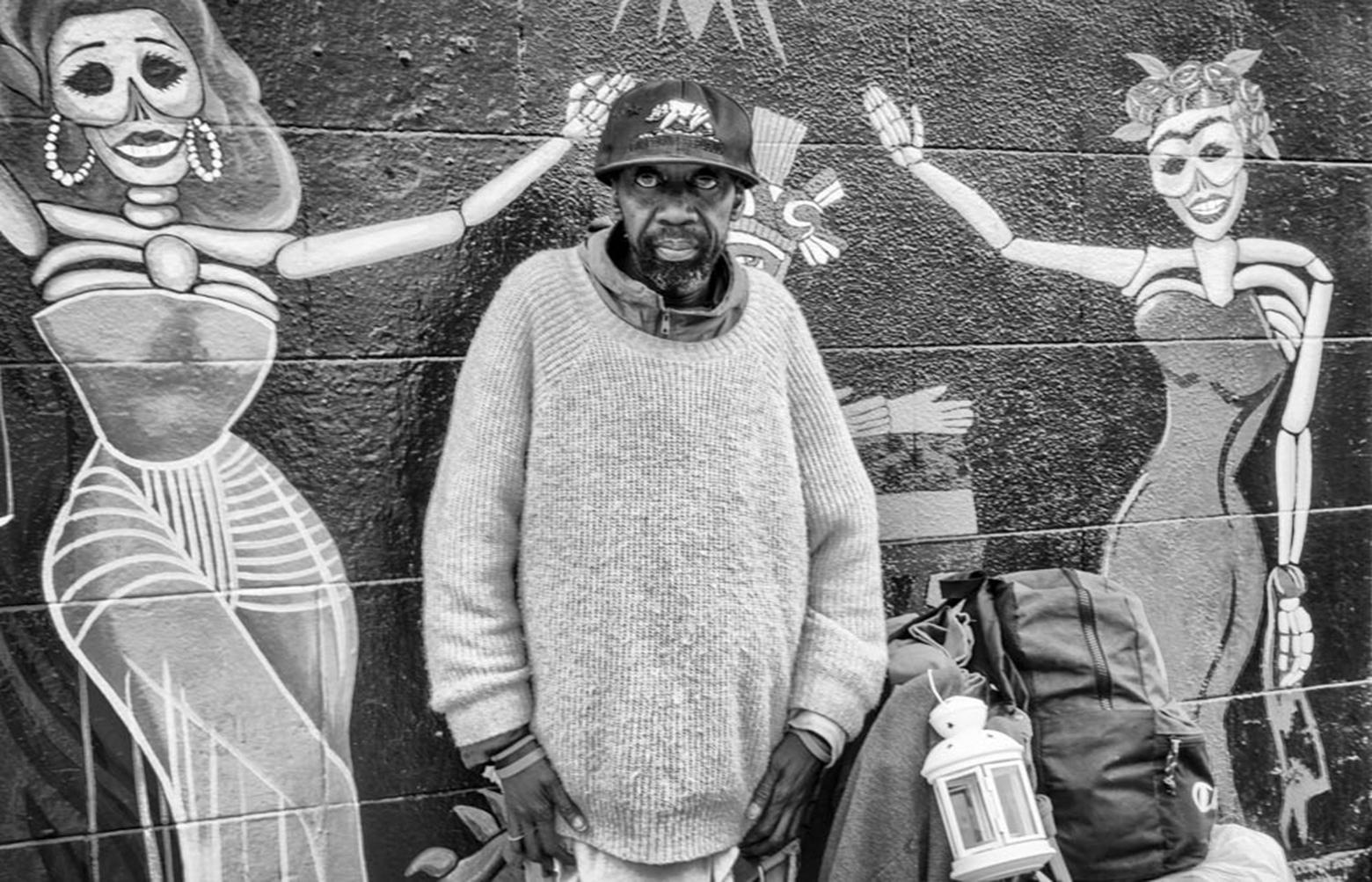 Abraham stands in front of a wall with a large mural of two masked party-going women. He is looking into the camera and is wearing a sweater and cap.
