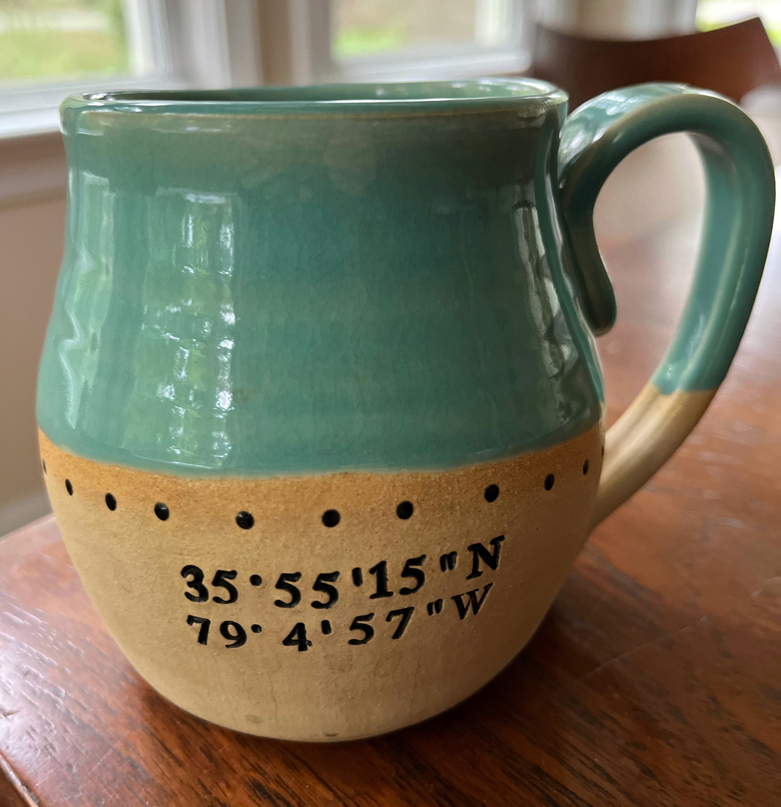 A coffee mug on a table in front of a window. On the lower half of the mug is a set of longitude and latitude coordinates.