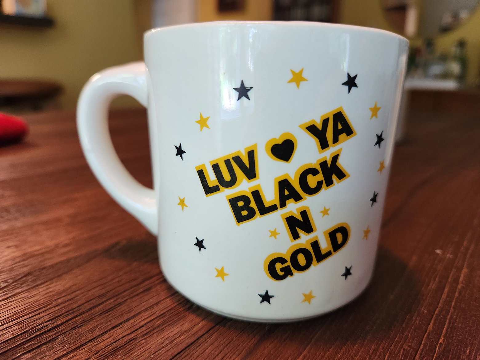 A white coffee mug on a table that says Luv Ya Black N Gold.
