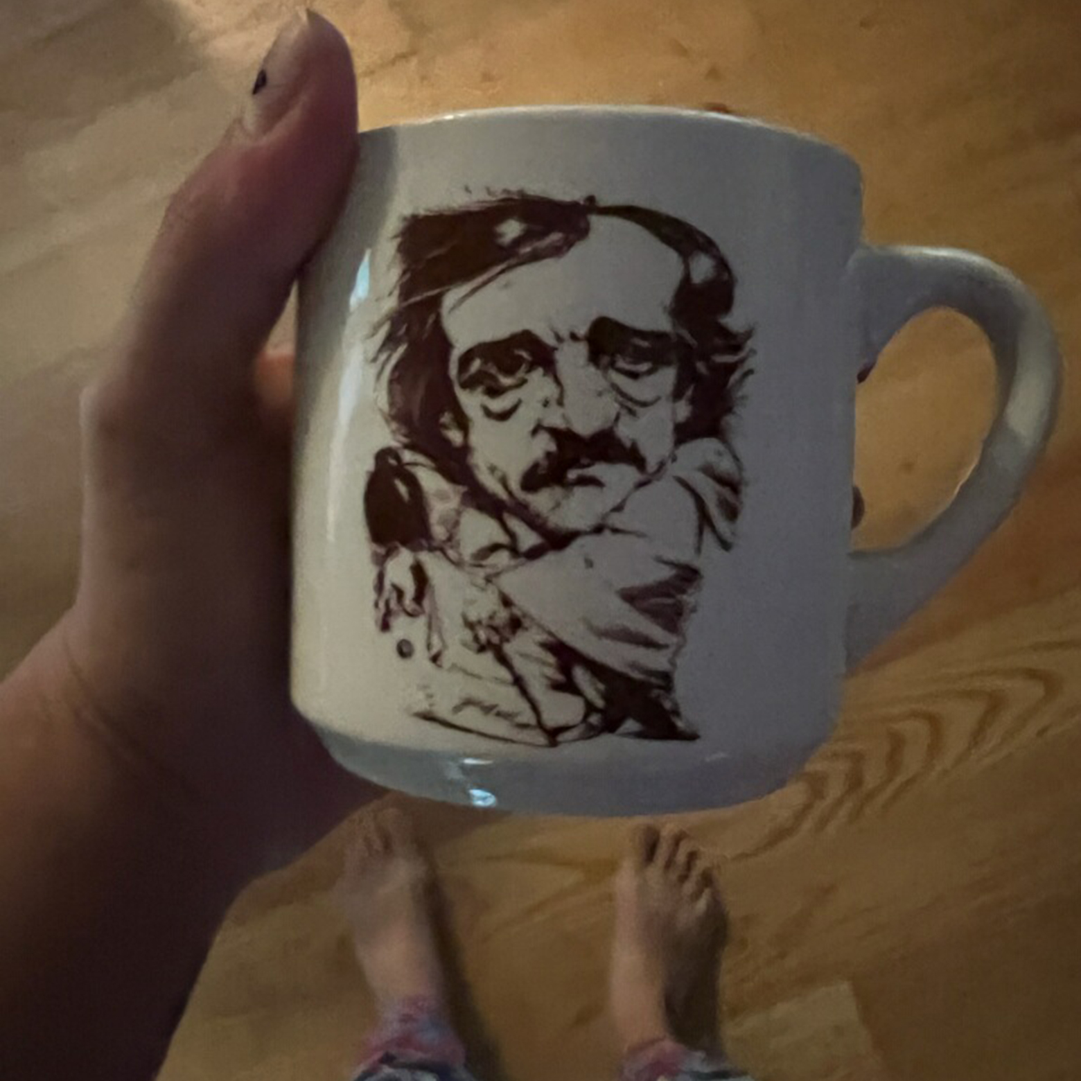 A white coffee mug is held up for the photo. The mug has a caricature of Edgar Allan Poe on it.