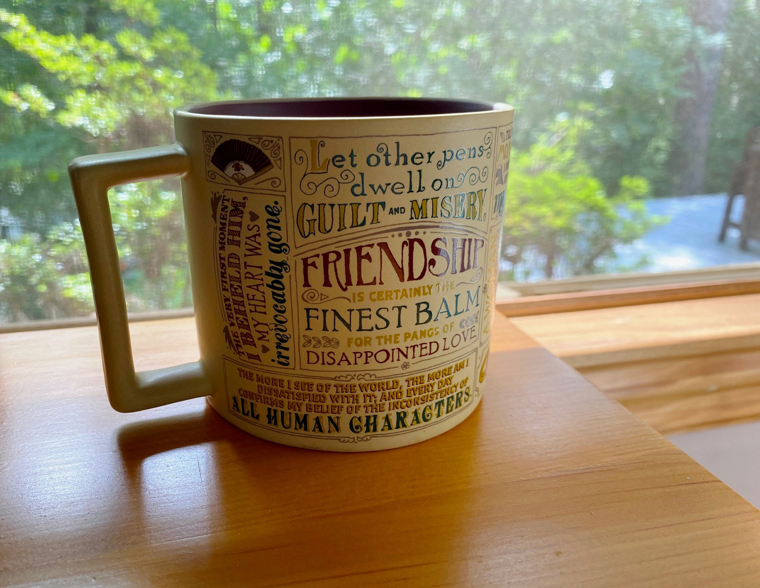 A coffee mug on a table in front of a window. The mug has quotes of Jane Austen in many sizes and styles.