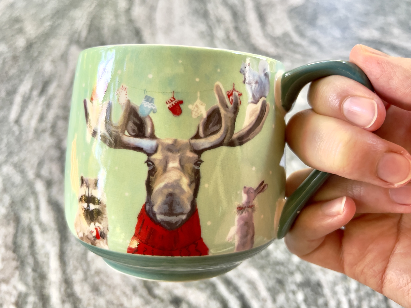 Christmas coffee mug is held above a countertop. The mug has a moose with a cardinal on the left antler and a squirrel on the right, each holding the end of a string with mittens hanging from it. A raccoon and a bunny flank the moose on the bottom.
