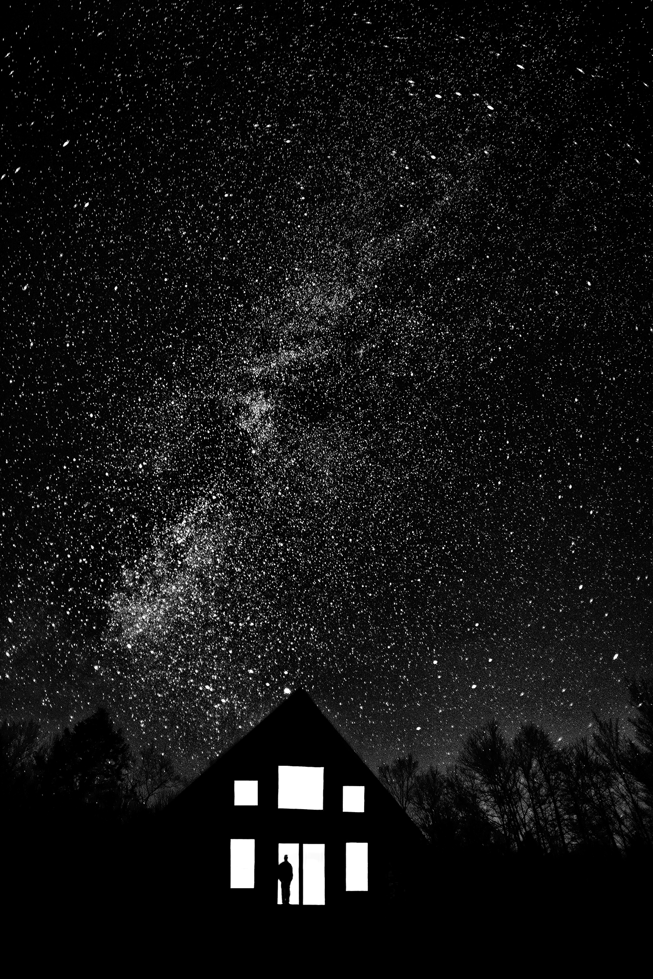 A man stands at the sliding glass door of a house in rural Pennsylvania looking out on a dark night at a sky blanketed with stars. The pitched-roof house is black against the tree line except for the illumination from the sliding door and windows.