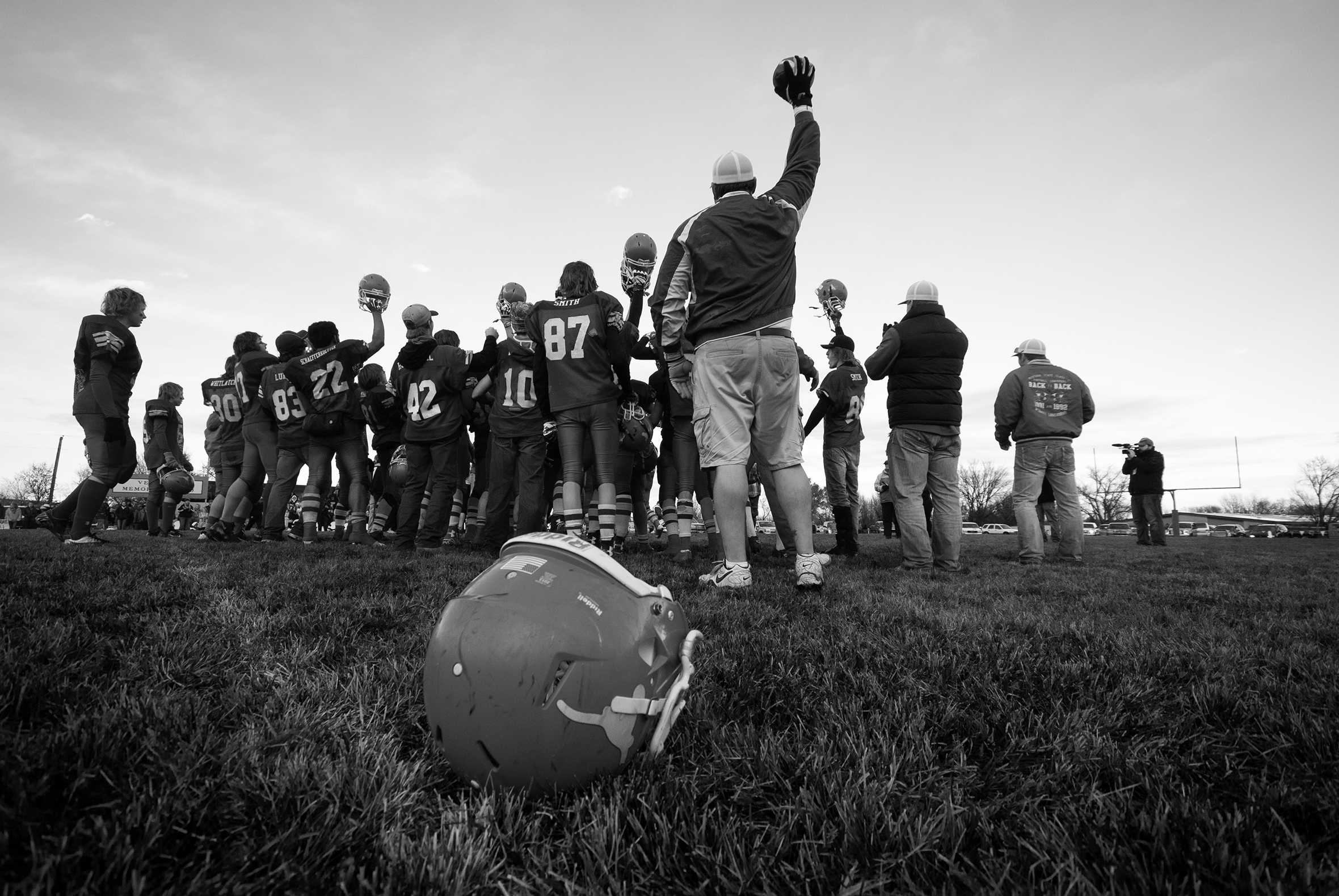 A school football team on the sideline of a field standing together with the coaches. One coach and several team members have an arm raised holding a football helmet in the air while a helmet rests on the grass in the foreground.