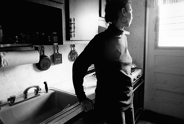 A woman stands with her back against the counter in a kitchen by the sink with her head turned to the left looking at the closed door.