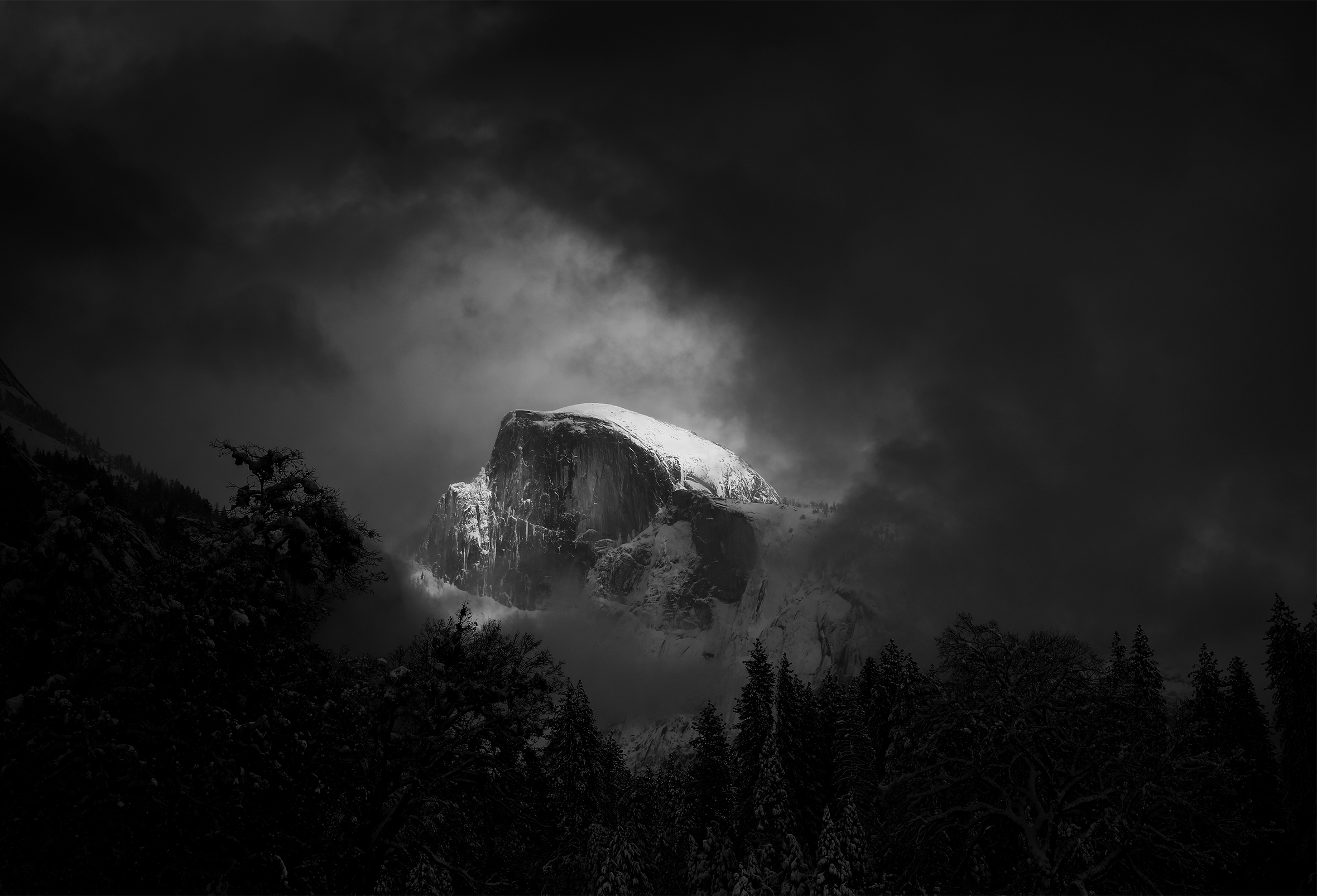 Treetops compose the foreground and provide framing for the sheer face of Half Dome at Yosemite National Park in California. Dark storm clouds surround the rock formation and fill the sky.