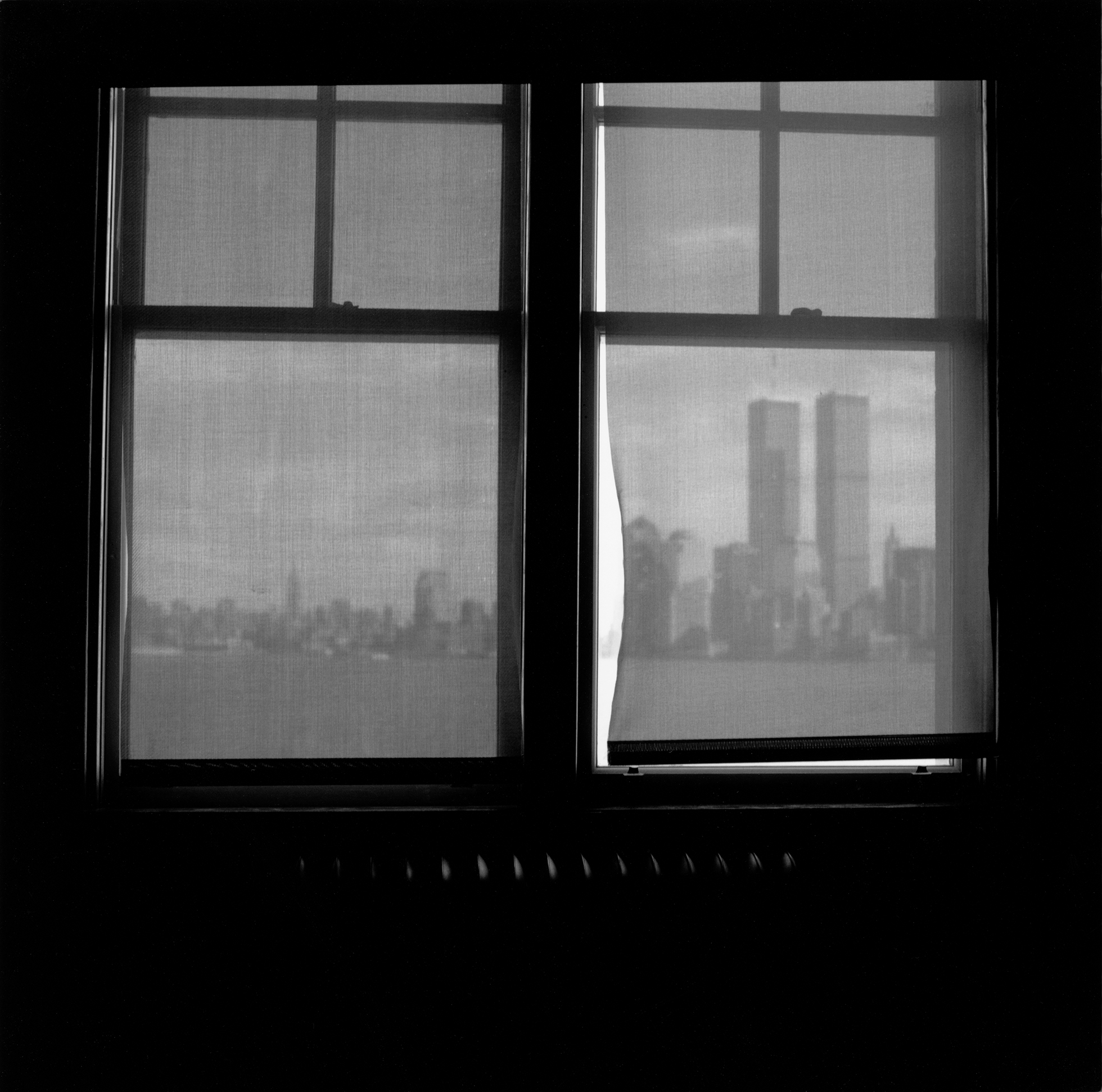 The New York skyline seen through twin windows covered with sheer shades with the Twin Towers visible in the right window.