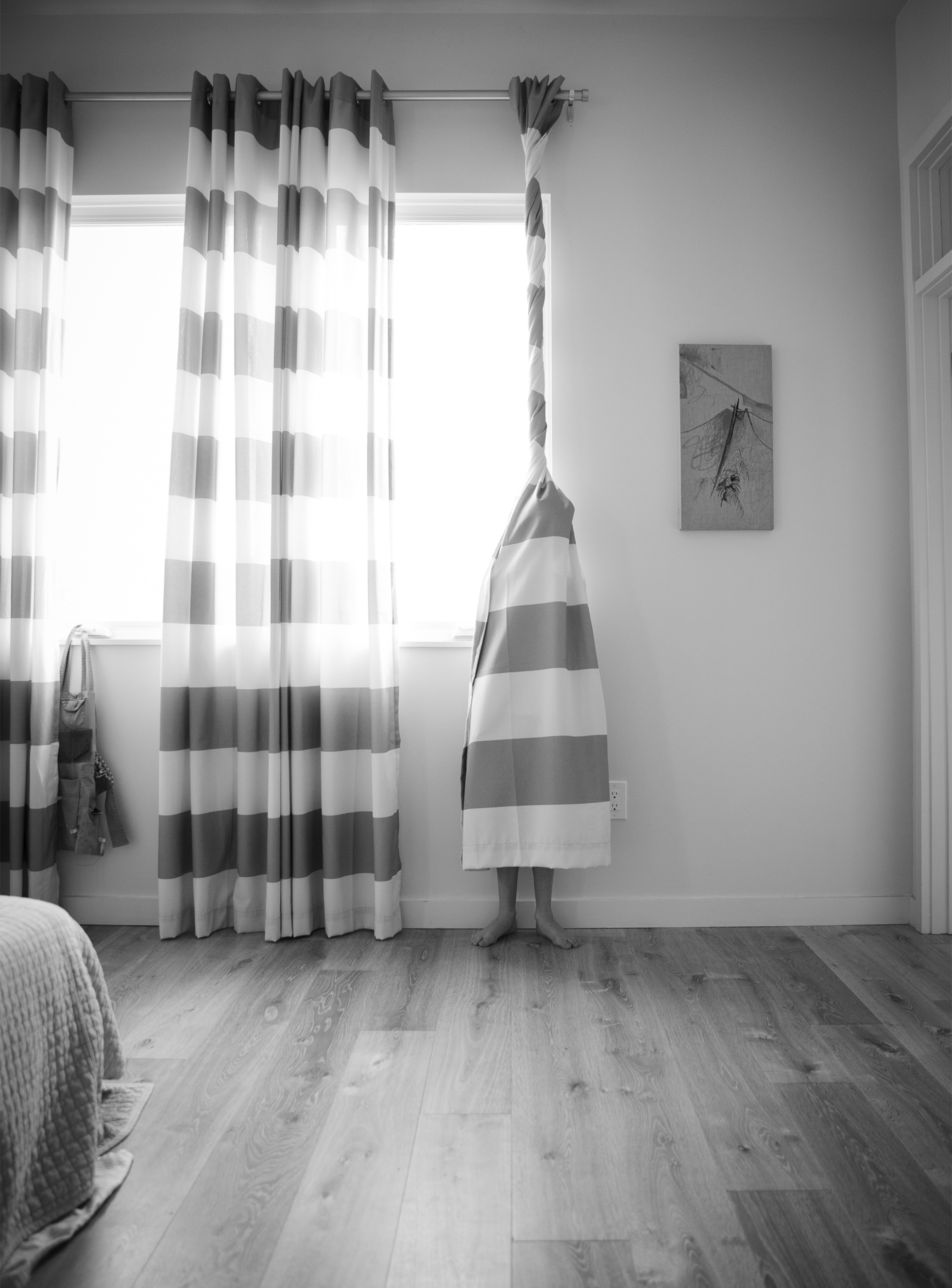 A child is wrapped in the right panel of a curtain with large horizonal stripes. The top of the curtain above the head is tightly wound and then the curtain flares out covering the body ending midcalf so part of the legs and bare feet are visible.
