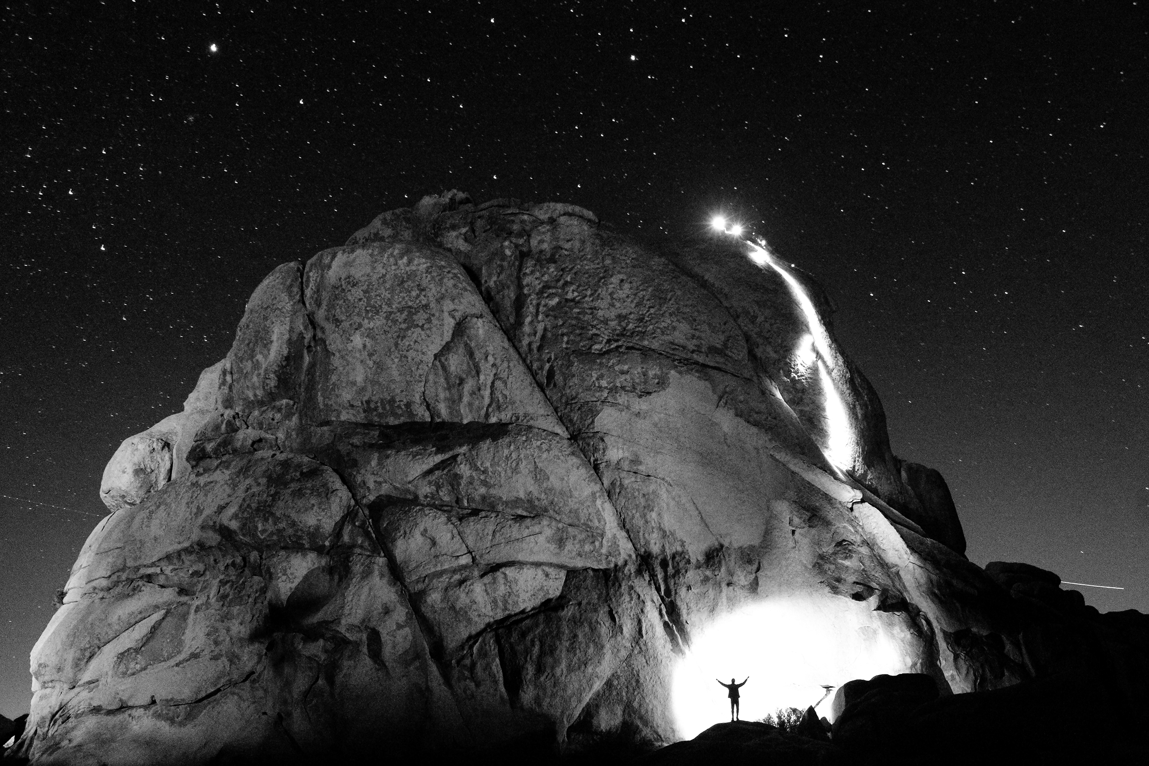 Two people rappel off a massive boulder in the desert of Joshua Tree National Park in Southern California at night under a blanket of stars while another person with outstretched arms cheers from the base. Spotlights illuminate the small figures.