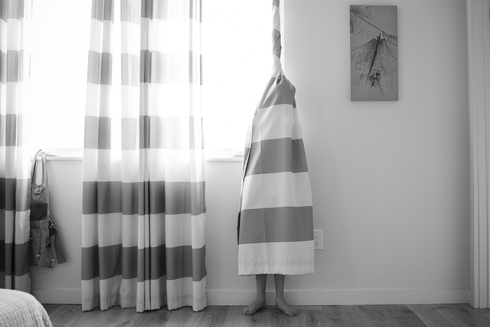A child is wrapped in the right panel of a curtain with large horizonal stripes. The top of the curtain above the head is tightly wound and then the curtain flares out covering the body ending midcalf so part of the legs and bare feet are visible.