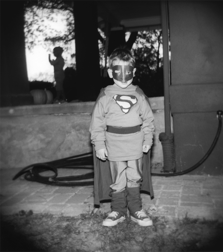 A young boy in a Superman costume with cape and mask stands facing the camera near the patio of a house.