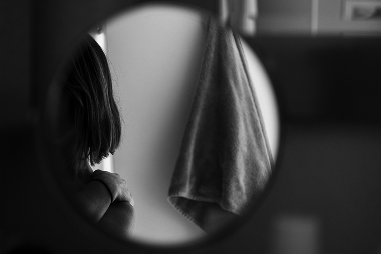 A glimpse of a woman with bobbed hair near a towel hanging from a hook on the wall seen in a circular bathroom mirror. The area outside of the mirror is blurry and dark.