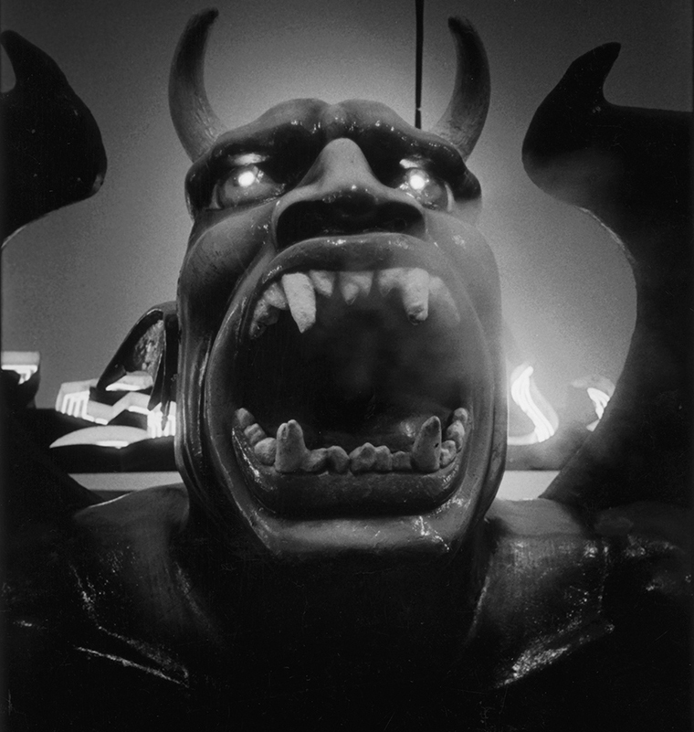 Closeup of a large figure with horns on the top of its huge head as its mouth is opened wide showing large teeth.