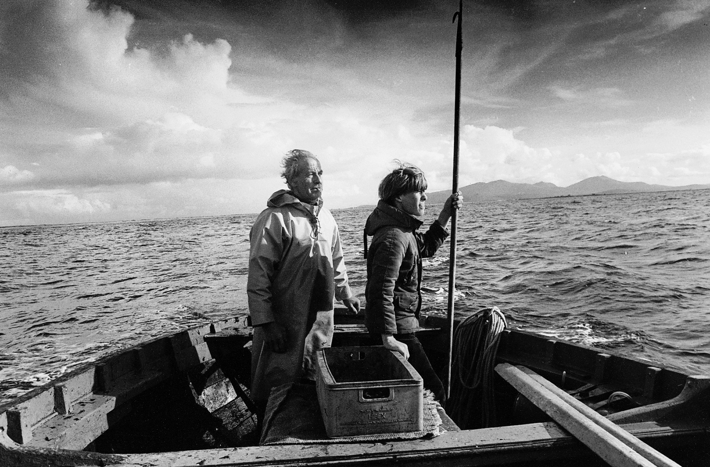 An old man and a young man both in profile looking off to the right in a nondescript boat off the Outer Hebrides, Scotland, on a choppy sea on a cold, cloudy day. The young man is holding a long-handled harpoon pointed up in his left hand.