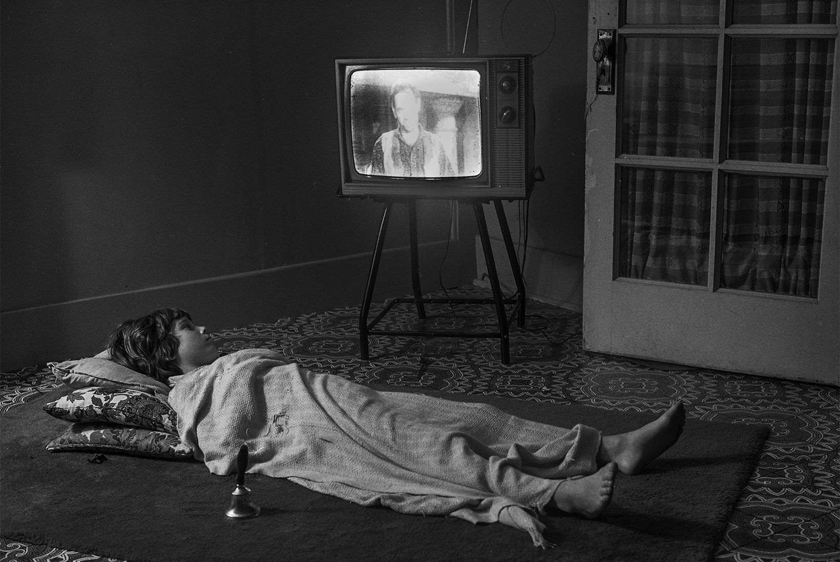 A young boy in the 1970s is flat on his back on the floor atop an area rug on top of carpeting with three pillows under his head and covered with a blanket in a room with an old-fashioned television. There is a long-handled bell near him on the rug.