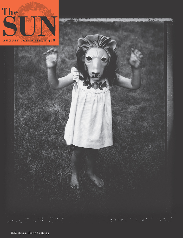 A young girl wearing a lion mask stands barefoot on grass in a white dress with her arms up bent at the elbows, palms facing forward, fingers curled like she is gesturing Roar!