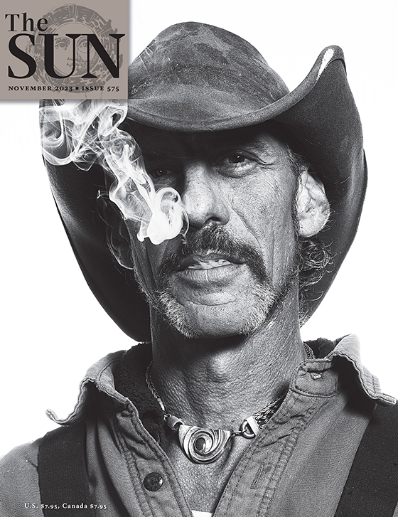 November 2023 cover of The Sun. A photo of artist, musician, carpenter, and mystic Doug Lipper taken while documenting his bond with a wild raven he raised. Sporting a cowboy hat and chunky necklace, Lipper has a plume of smoke coming from his mouth.