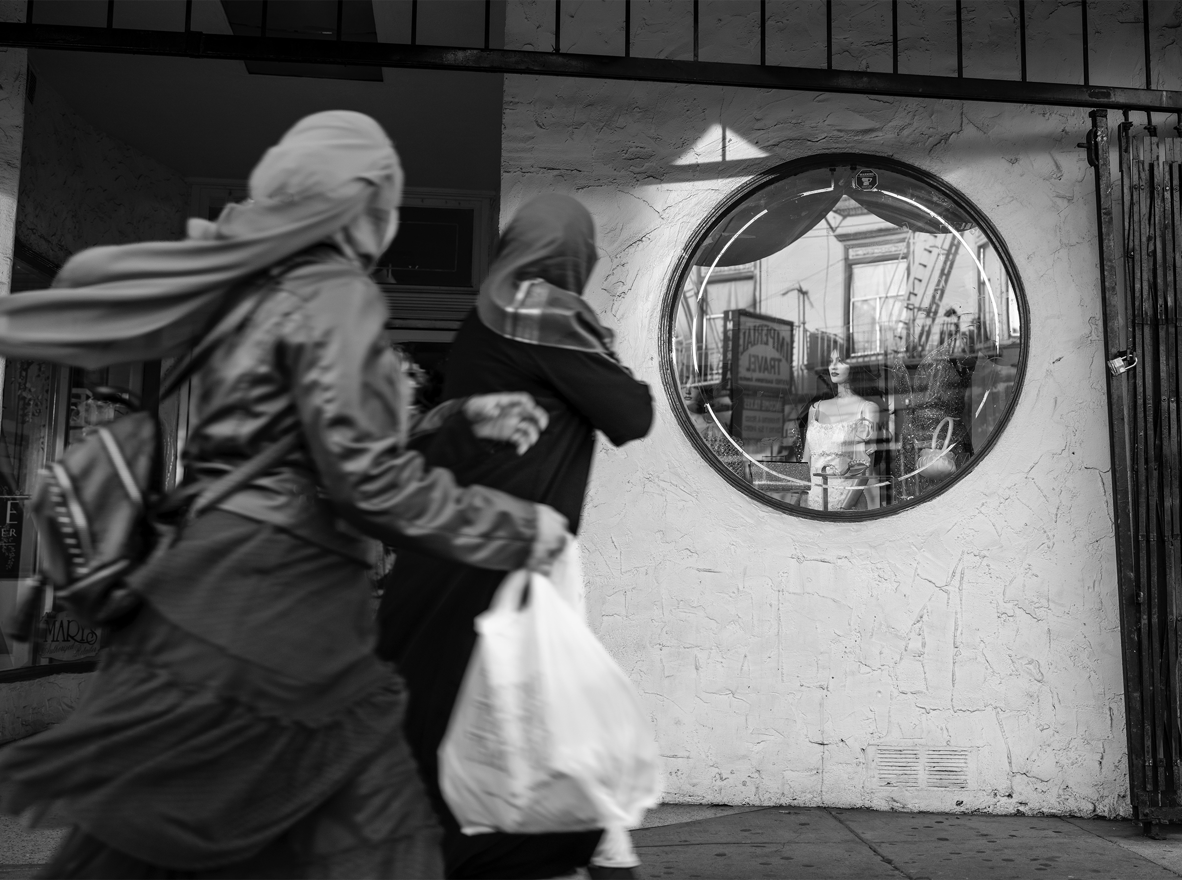 Two women in headscarves window-shop on Mission Boulevard in San Francisco. They walk by a women’s clothing store with a circular window displaying evening purses, strappy sandals, and mannequins in dresses.