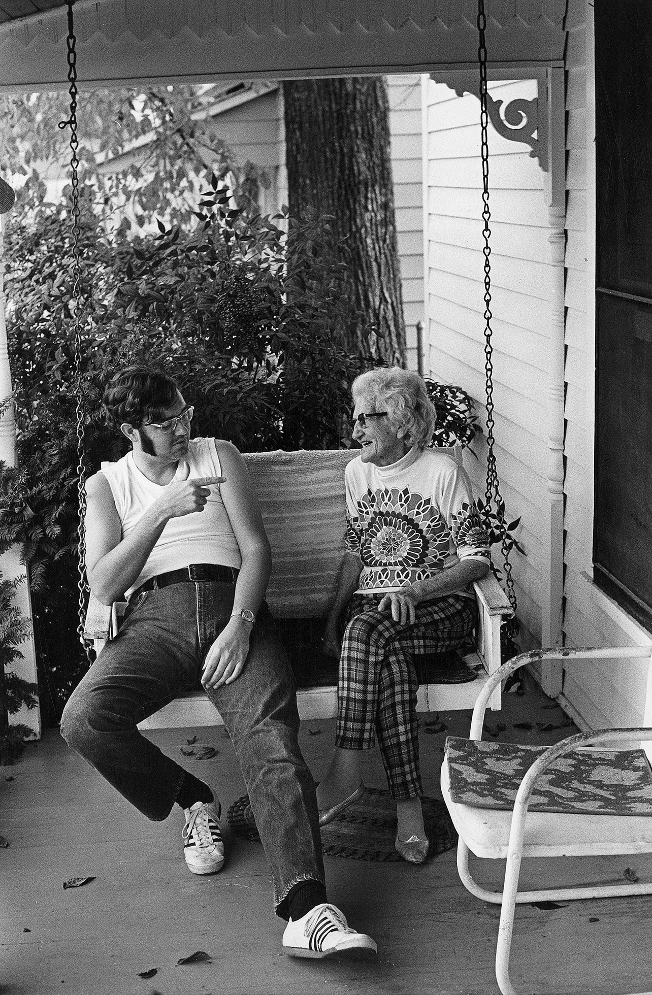 A young man with long, thick sideburns and glasses wearing a white sleeveless T-shirt, jeans, and “track shoes” circa 1970 and an old woman in a sweater and plaid pants sit and talk on a suspended swing on a porch. He points at her while she smiles.