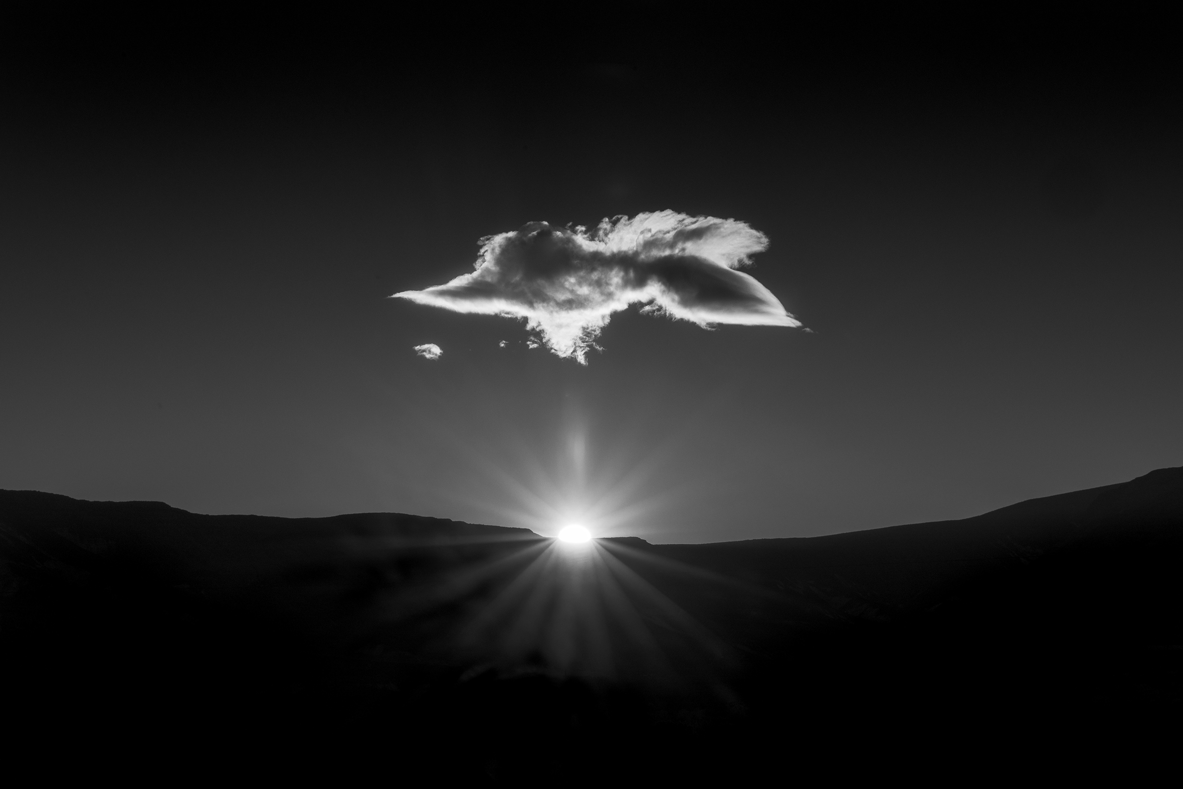 Bright sun with prominent rays rises up over a hilly, barren landscape with only one cloud in the sky which is perfectly positioned and illuminated directly above the sun.