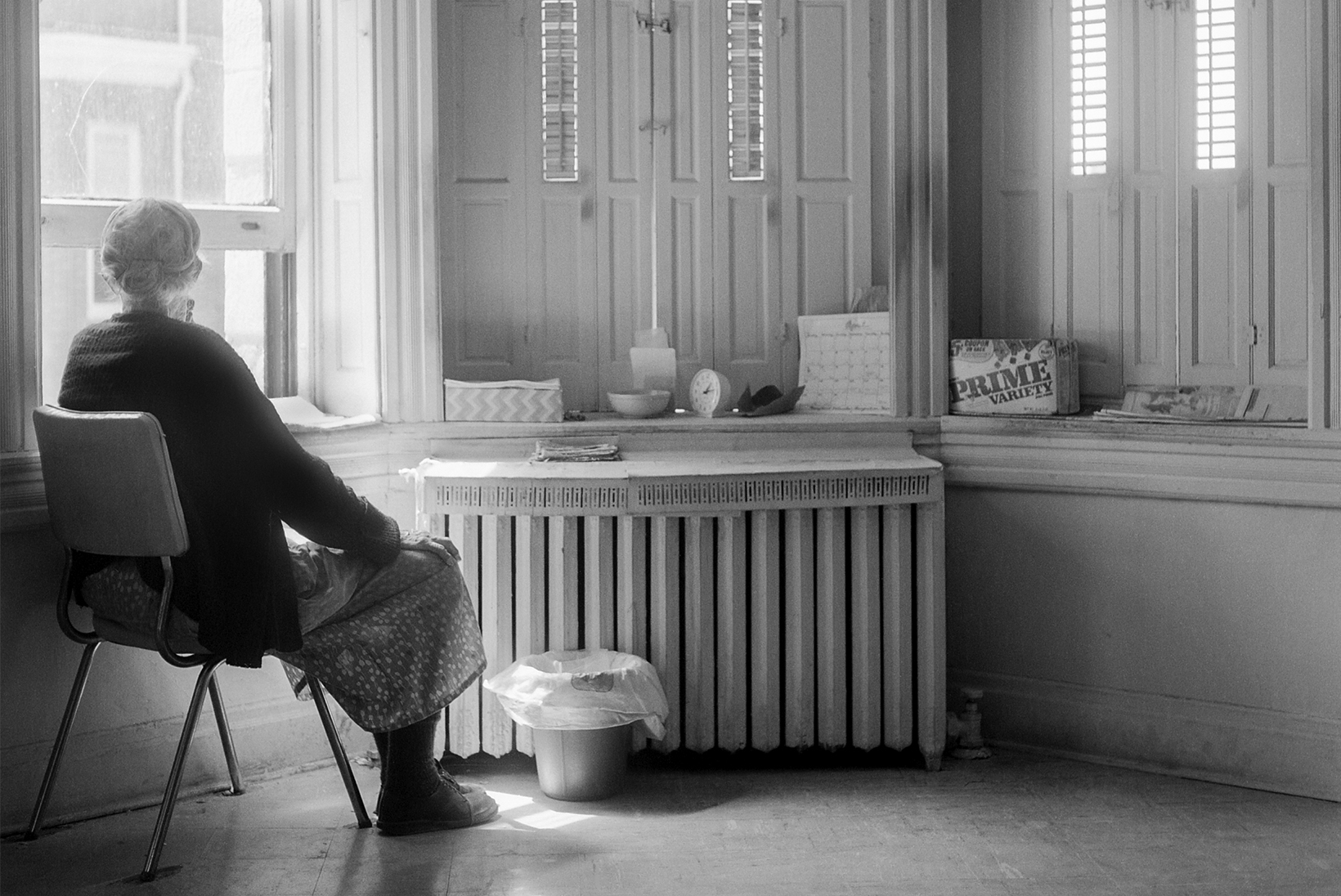 An old woman, with her hair in a bun and wearing a dress and sweater, sits in a chair and looks out of the window. The room has a steam radiator air vent, tall windows with interior shutters, a small wastebasket, and personal items on the windowsill.