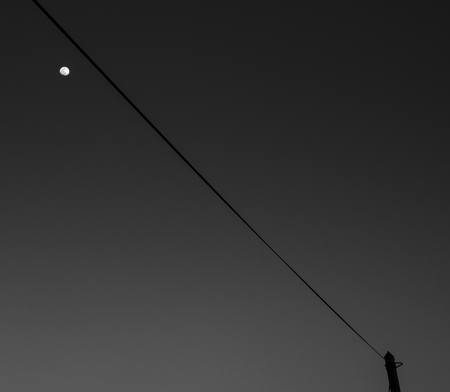 A telephone wire traverses the sky from top left to bottom right where the top of a telephone poll peeks up. The only object in the night sky is a tiny full moon under the telephone wire in the upper-left corner.