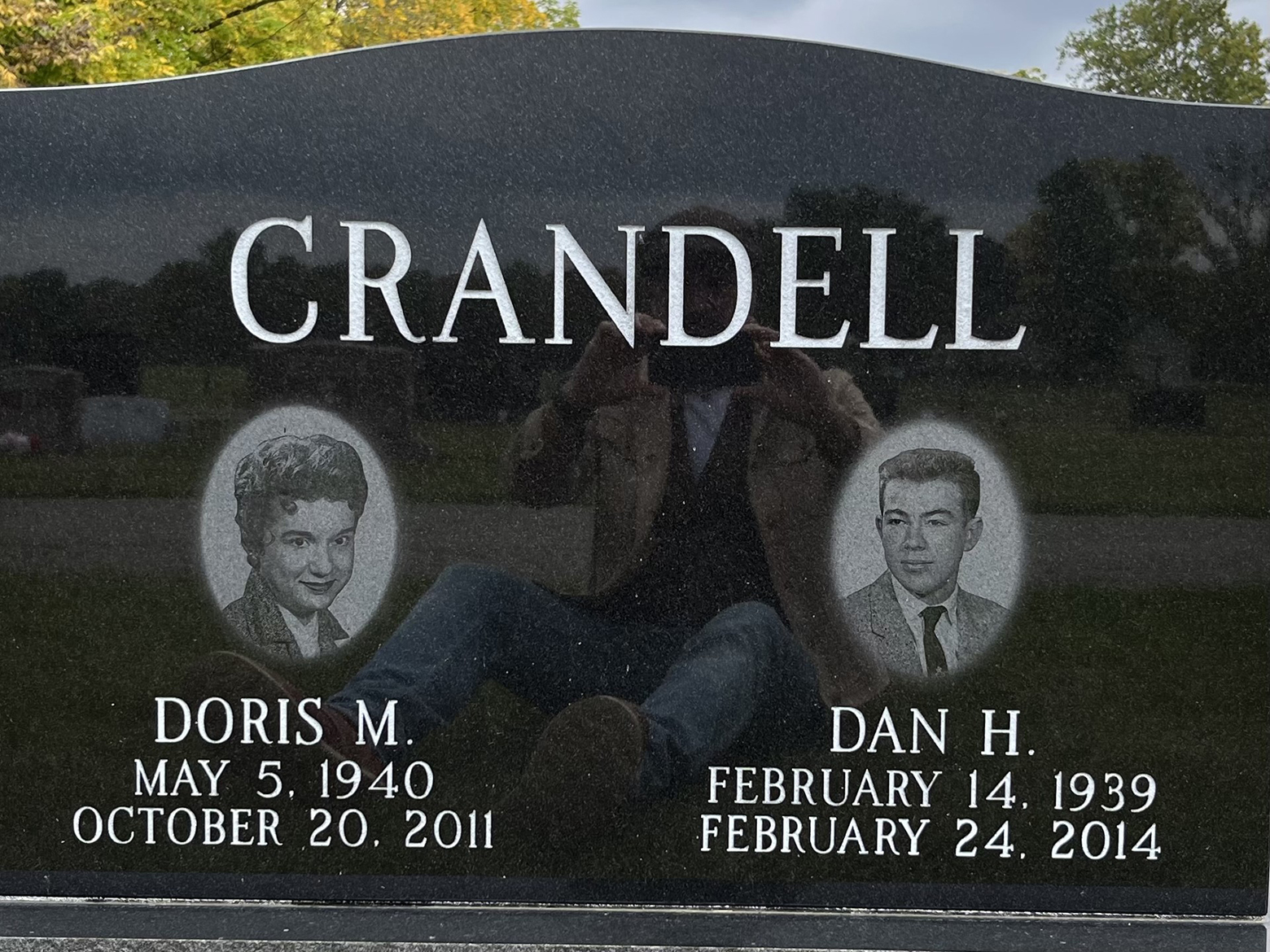 Doris M. Crandell and Dan H. Crandell’s headstone with images of them etched into it. Other headstones are reflected in the Crandell’s headstone. Also reflected is an image of Doug Crandell sitting down while photographing his parents' headstone.