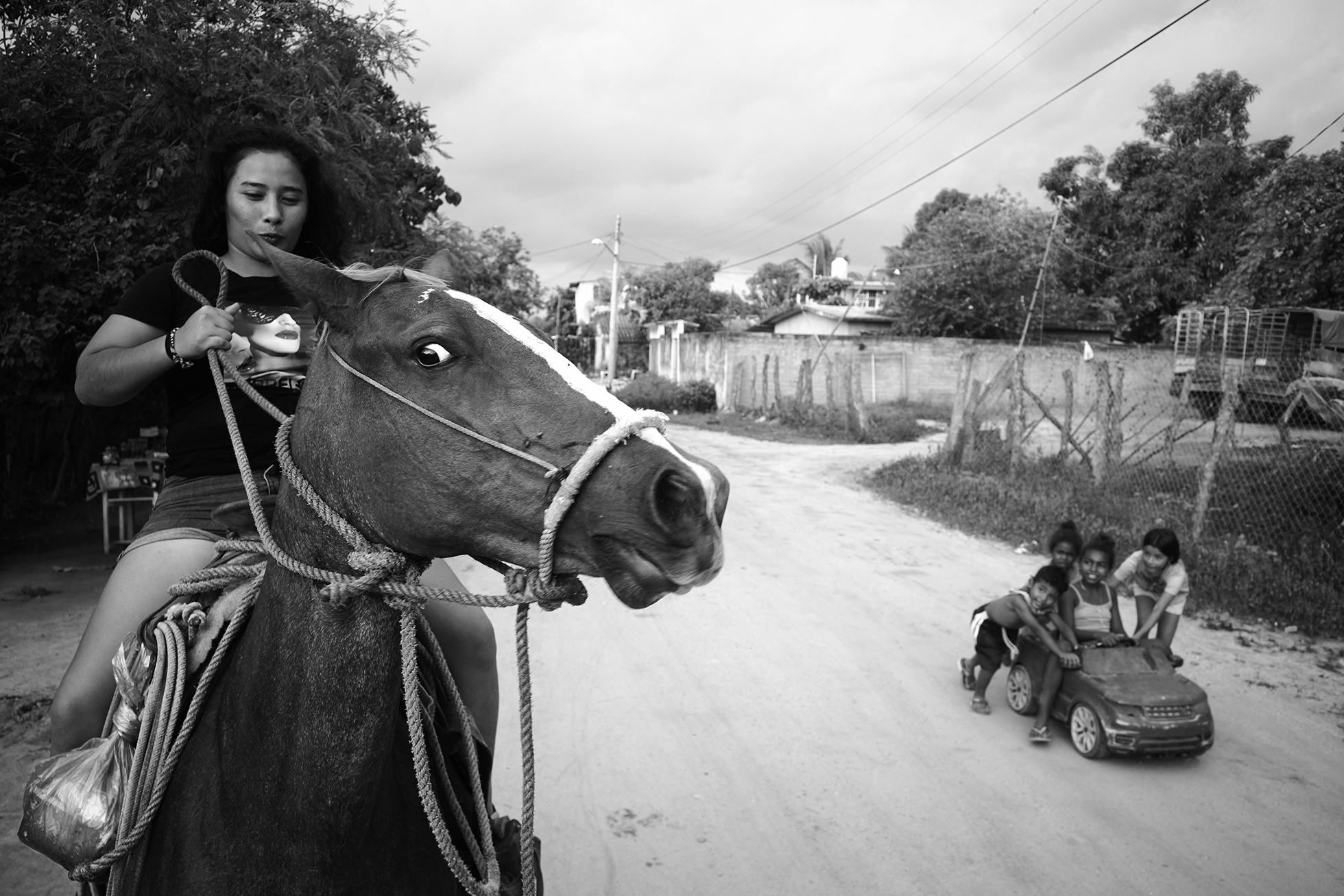 A woman rides a horse down a dirt road while four children in and around a ride-on toy car look on.