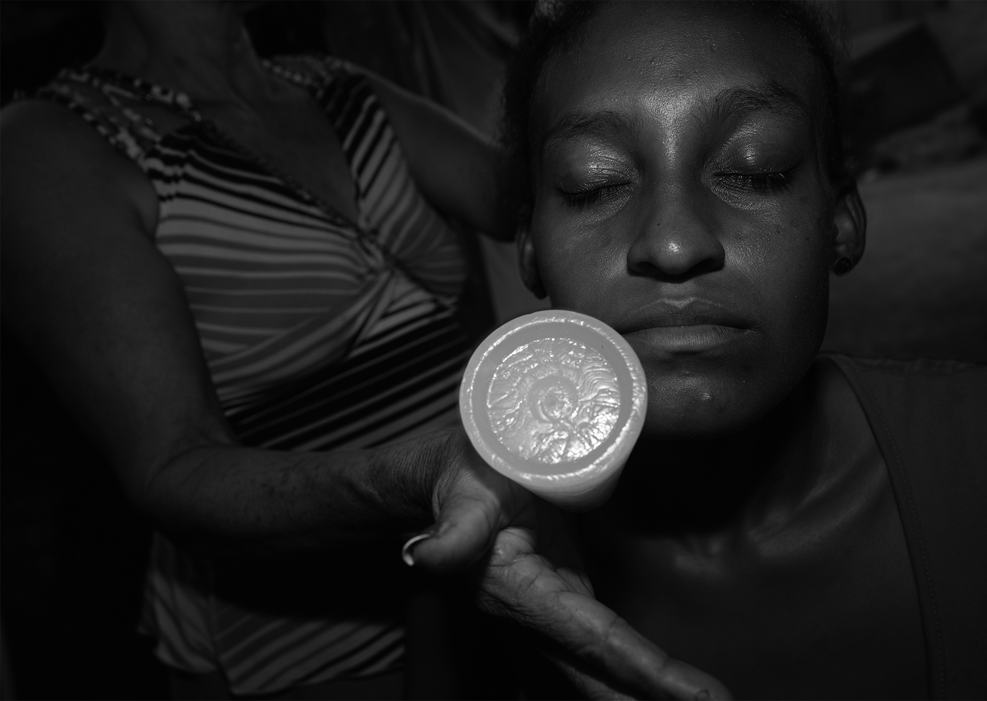 A young woman is treated for an illness by a traditional healer. The young woman closes her eyes as the healer rolls an object on the side of her face.