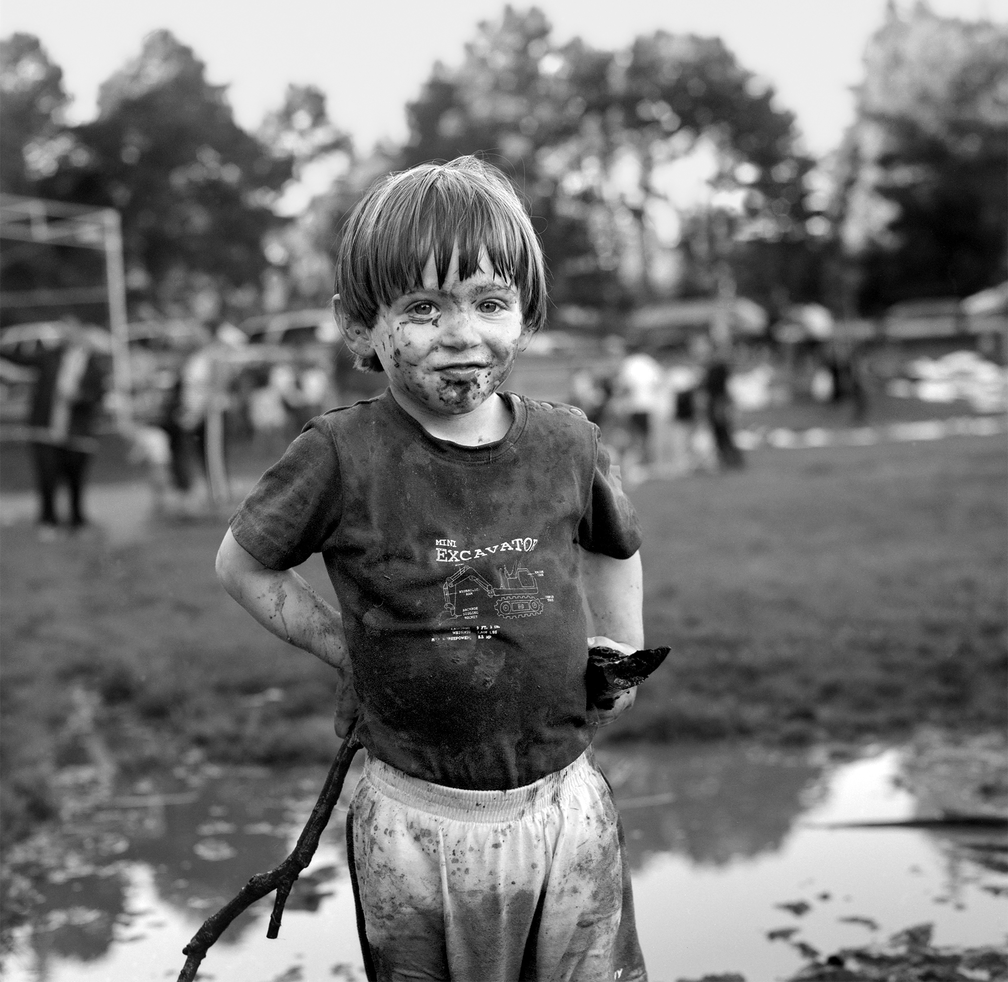 A small boy wearing a T-shirt and once-white shorts stands in a large puddle holding a stick as he looks at the camera in a public park in Oakland, California. He is covered in mud and looks pleased with himself.
