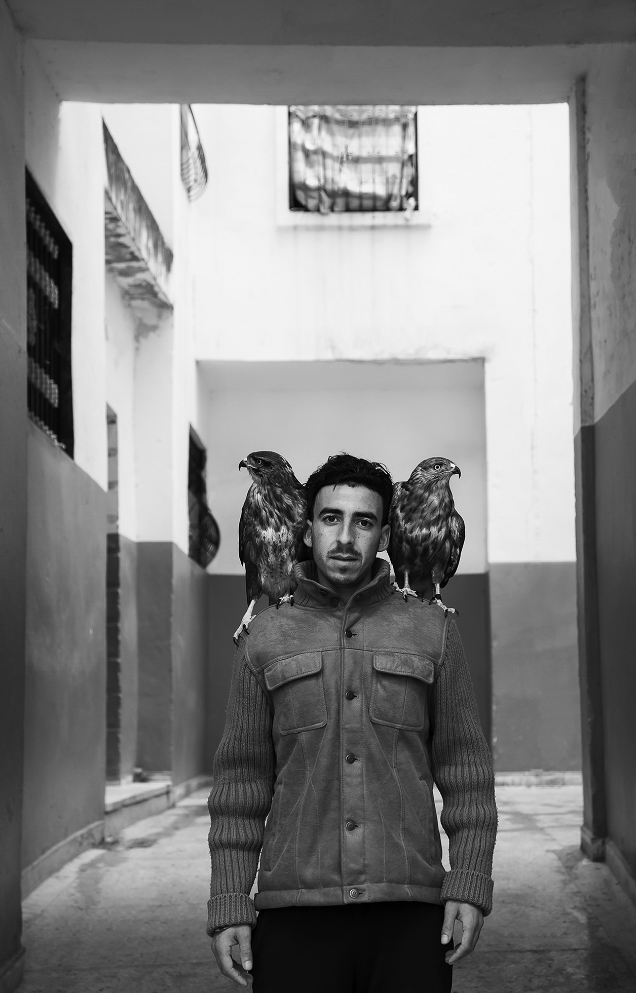 A falconer near his home in Tangier, Morocco, is wearing a long-sleeved jacket as he stands looking at the camera with a falcon perched on each shoulder: Diablo and Paco are each looking off to their respective side.