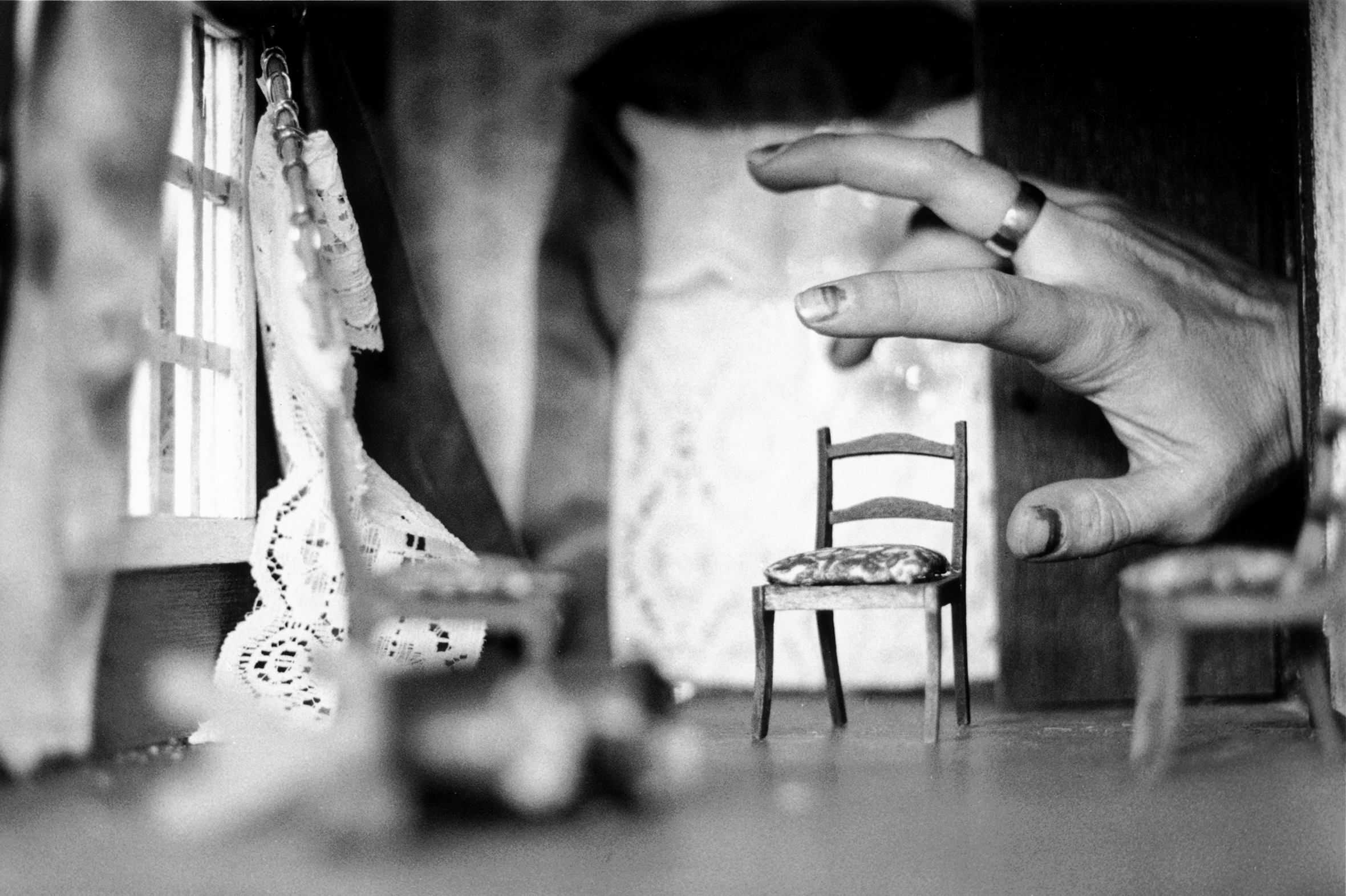 A woman's hand rearranges the furniture in an interior room in a dollhouse that has a window with curtains and three chairs.