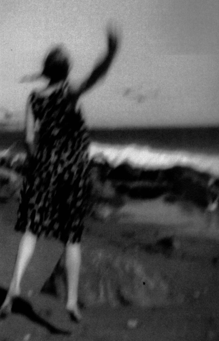 Blurry photo of a woman on the beach facing the water with her back to the camera. She is wearing a dress and is barefoot as she jumps up and waves her right hand with her arm fully extended.