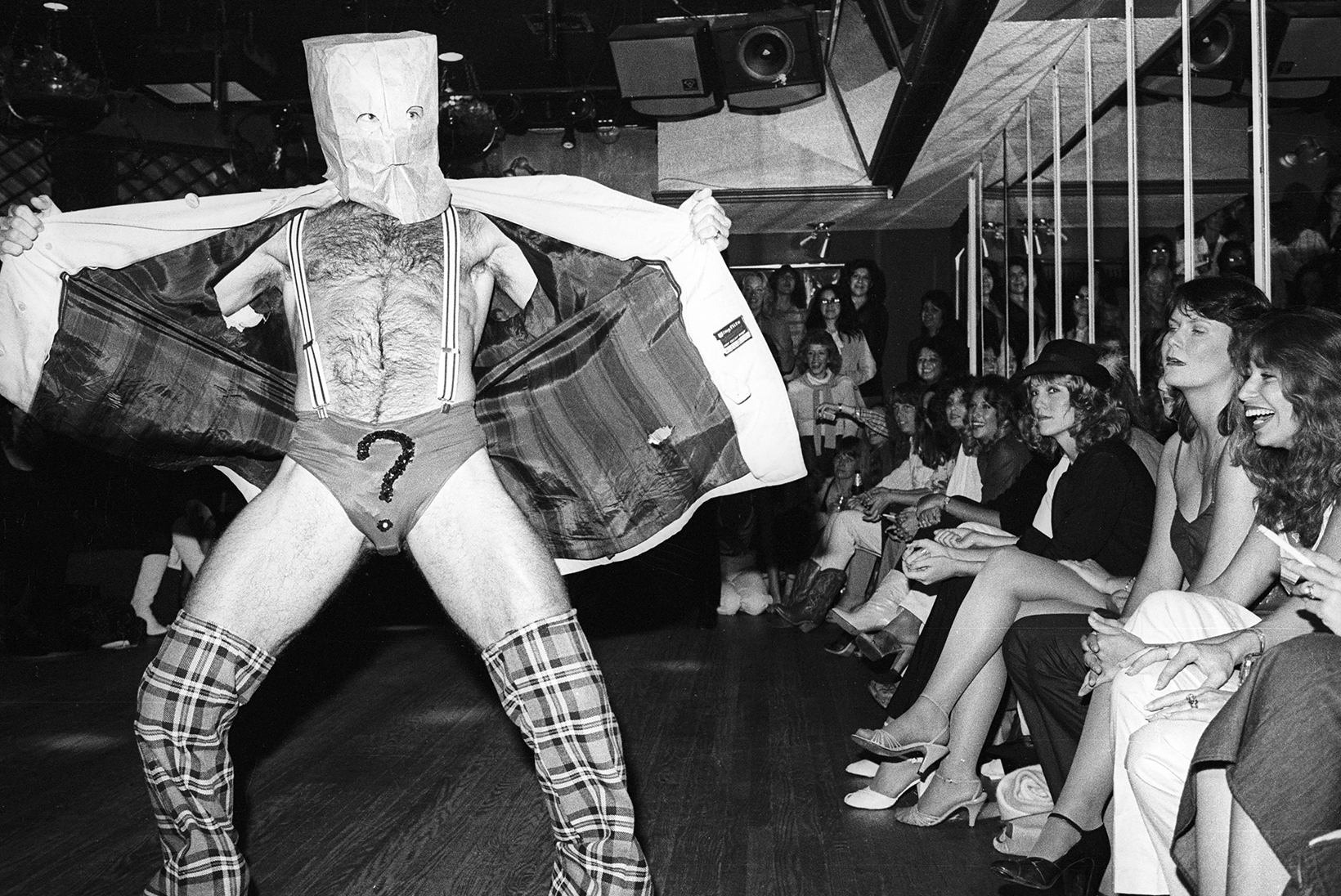 Chippendales dancer in Los Angeles in 1981 performs with a paper bag with cutouts over his head. He stands and opens a sports jacket revealing suspenders attached to underwear with a large question mark on the front.
