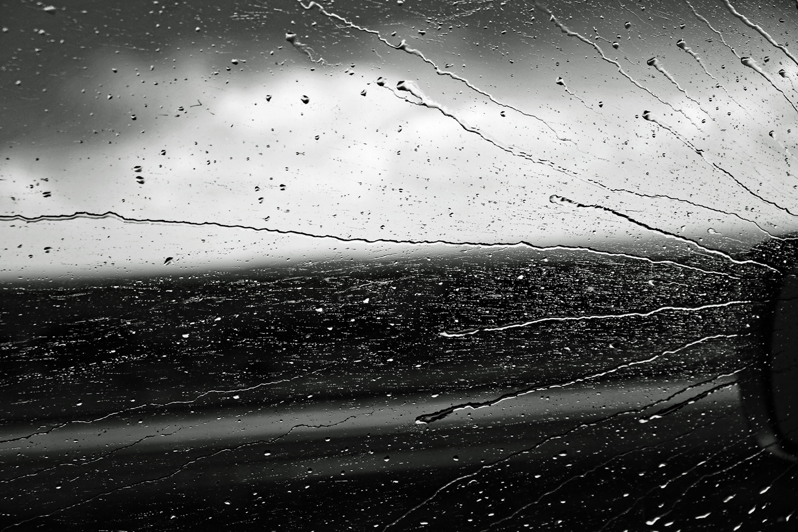 Rain streaks from right to left across a driver’s side car window viewed from inside a moving car. A small portion of the side mirror on the left is barely visible against the cloudy gray sky and the dark landscape.