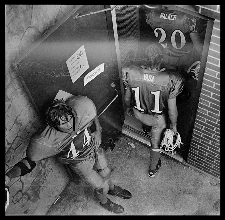 High-school football players walk into a building. One player stands with his back against the open door allowing his teammates to file in.